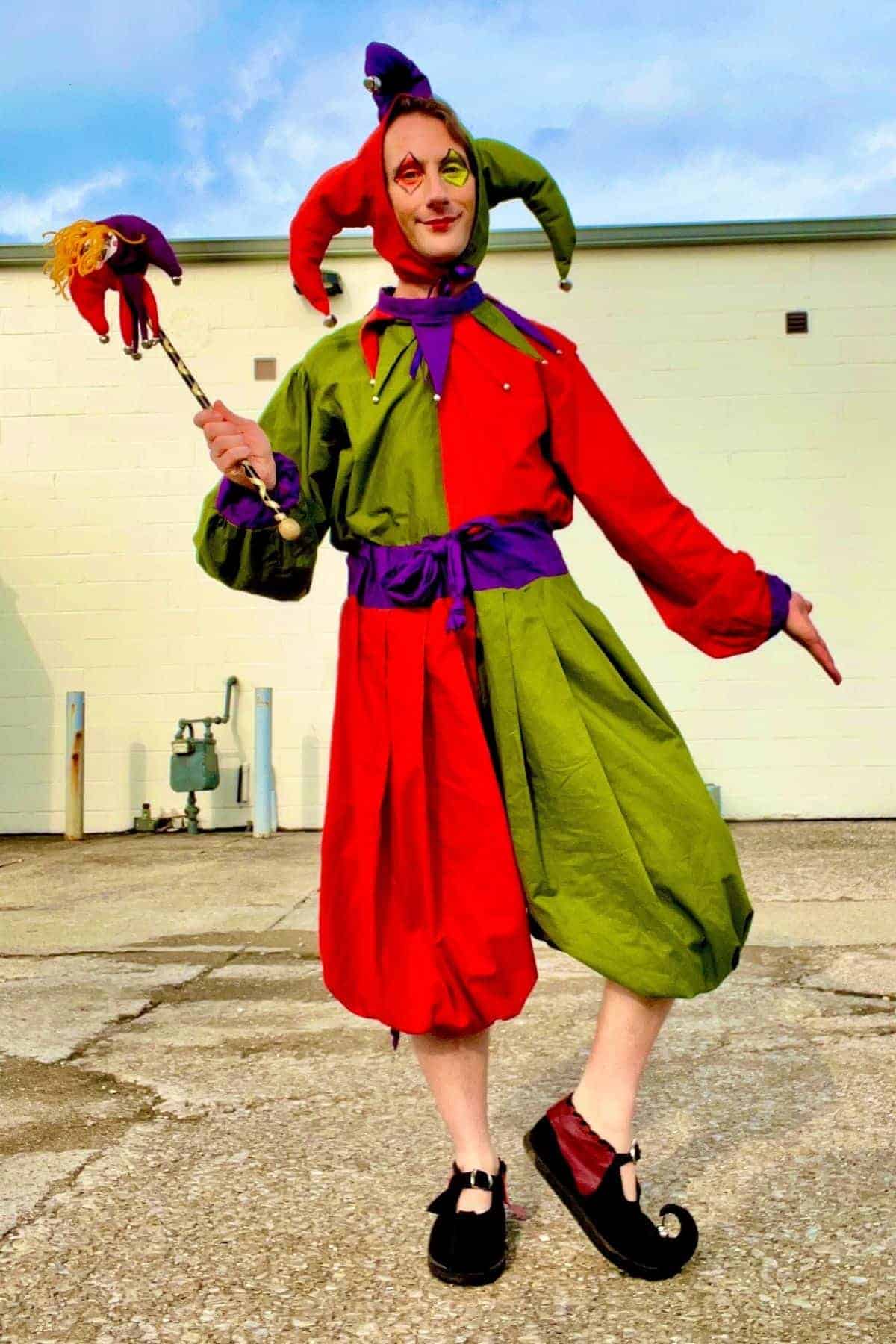 Person dressed as a jester in red and green costume, holding a matching jester stick, stands on pavement in front of a light-colored building.