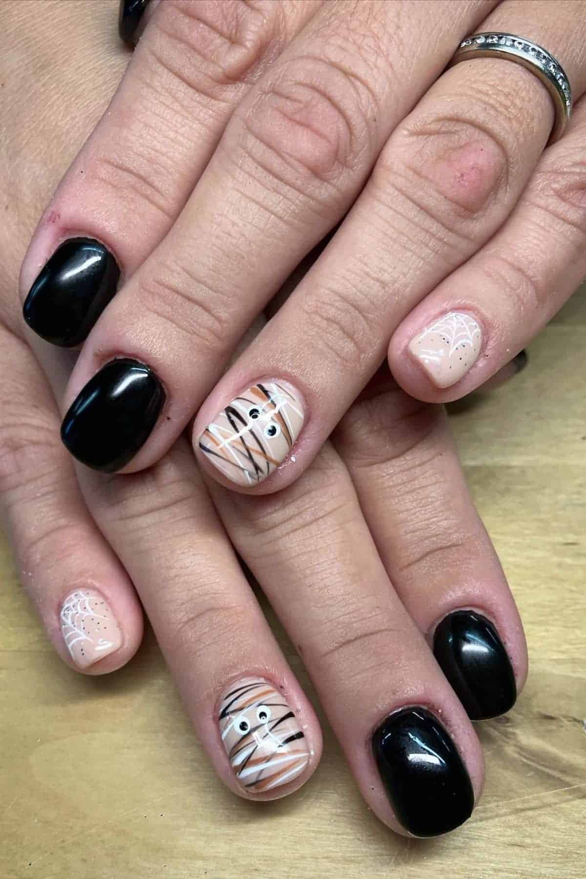 Hands with short nails painted black, except for accent nails with mummy and spider web designs in nude, white, and black, set against a light wooden surface.