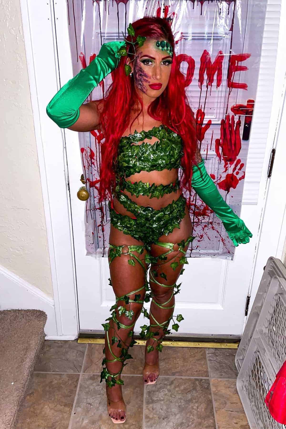 Woman in a leaf and vine costume with long red hair stands in front of a door decorated with fake blood handprints and “WELCOME” written in red.