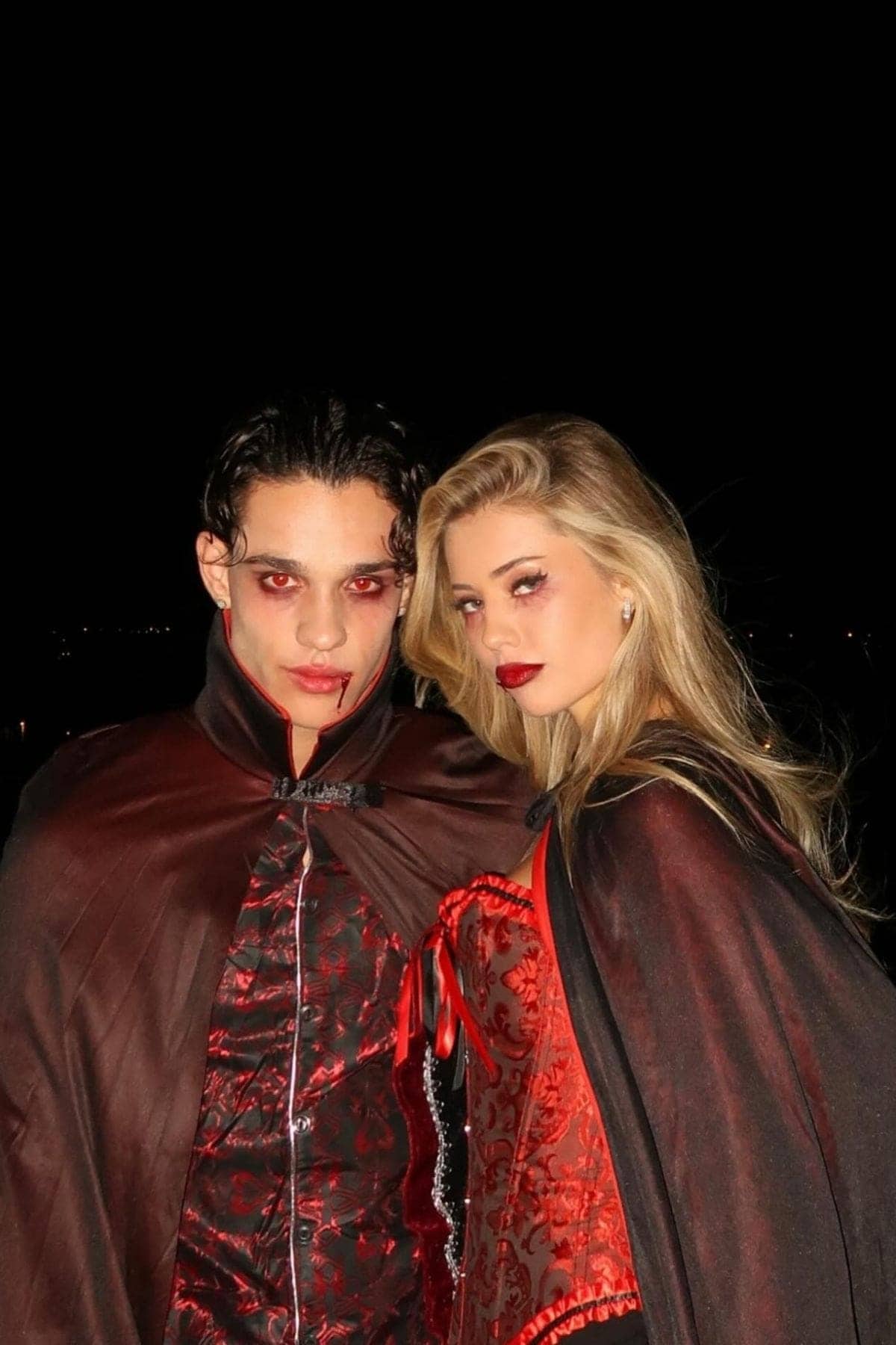 Two people in vampire costumes with red eyes, dramatic makeup, and black capes stand together against a dark background, gazing directly at the camera.