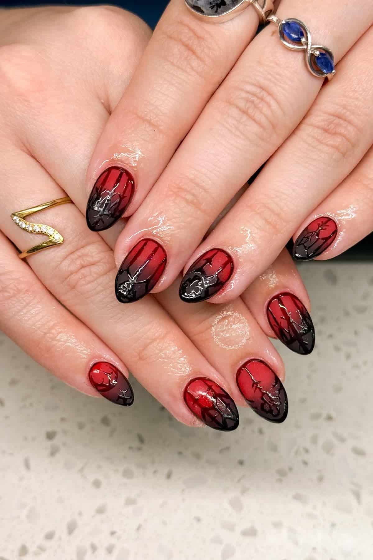 Hands with long, almond-shaped nails painted red with black and silver abstract designs. The person wears a gold zigzag ring and a silver ring with blue stones. White countertop background.