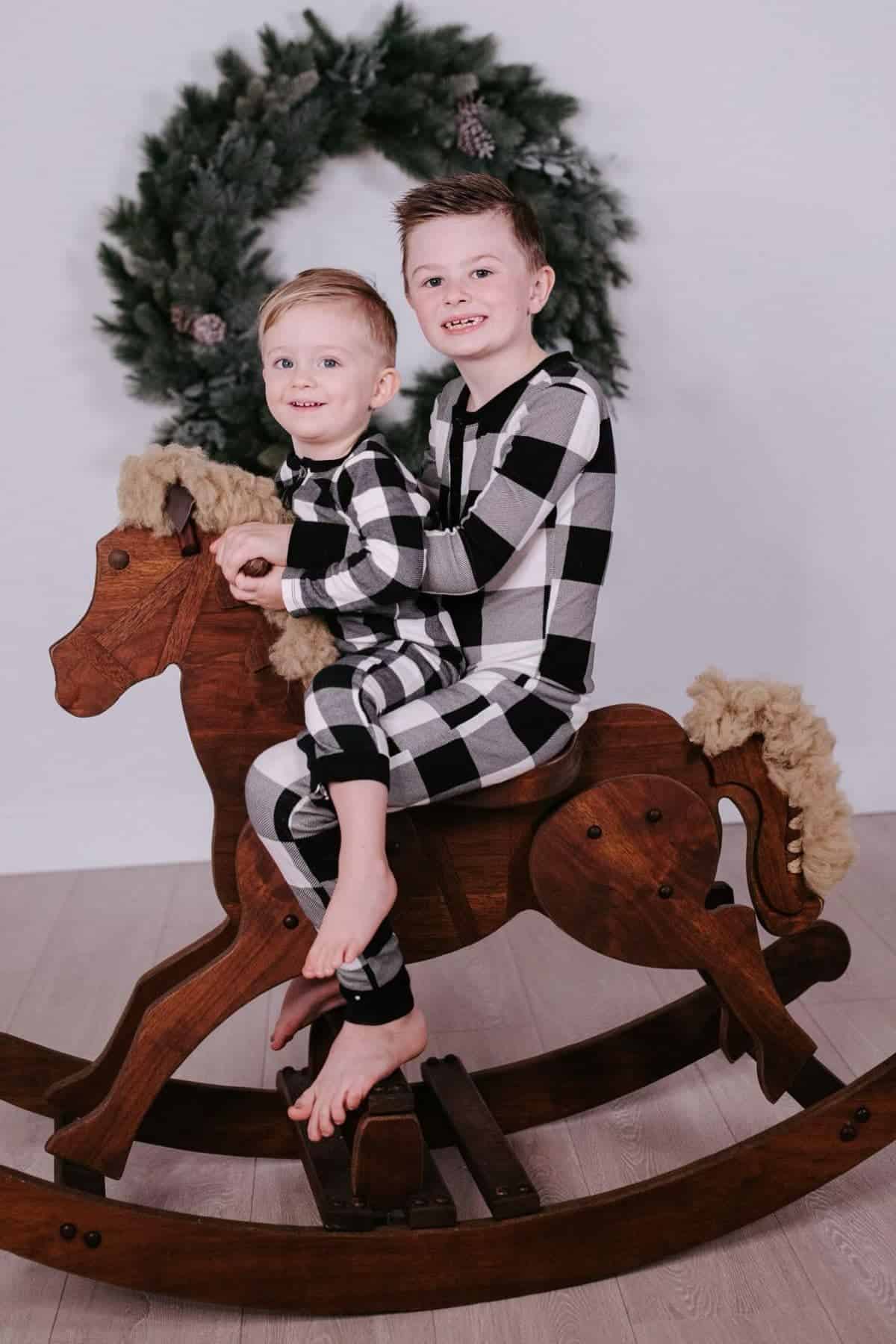 Two young children in matching plaid pajamas sit on a large wooden rocking horse with a green wreath hanging on the wall behind them.