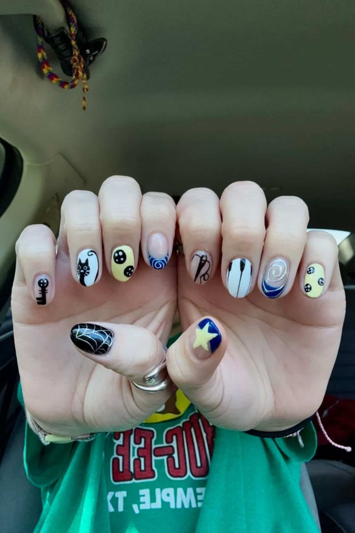 Two hands display nail art featuring cartoonish designs, including black cats, yellow smiley faces, swirls, a spiderweb, and a blue star, photographed inside a car.