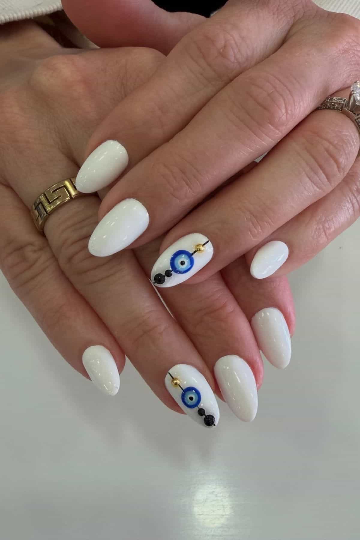 Hands with white, almond-shaped nails; one nail on each hand features an evil eye design with black dots and gold accents.