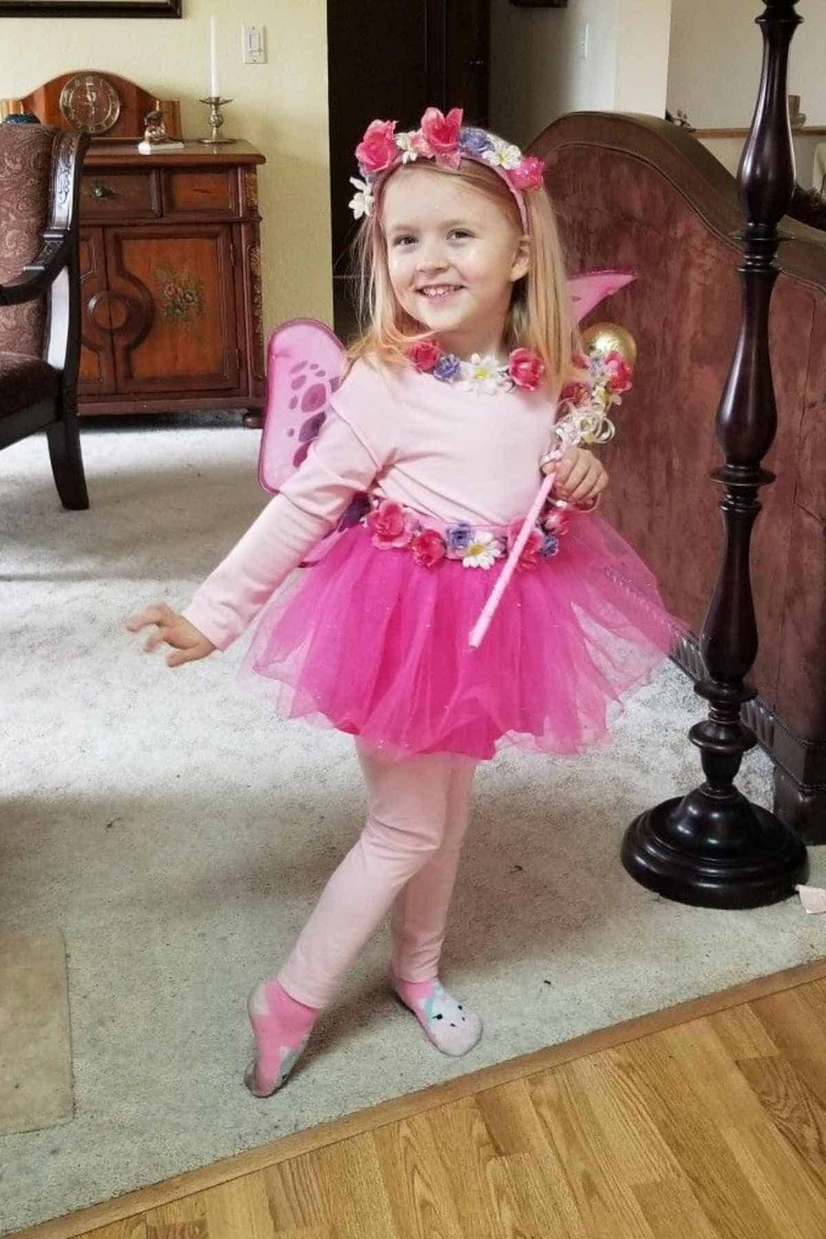 Young girl dressed as a pink fairy with wings, flower crown, tutu, and holding a wand, standing and smiling in a living room.