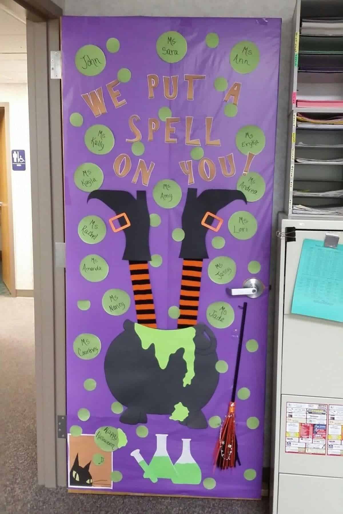 Decorated door with a witch’s legs in a cauldron, green dots, names on circles, and the phrase “We put a spell on you” in cut-out letters. Test tubes and broom also shown.