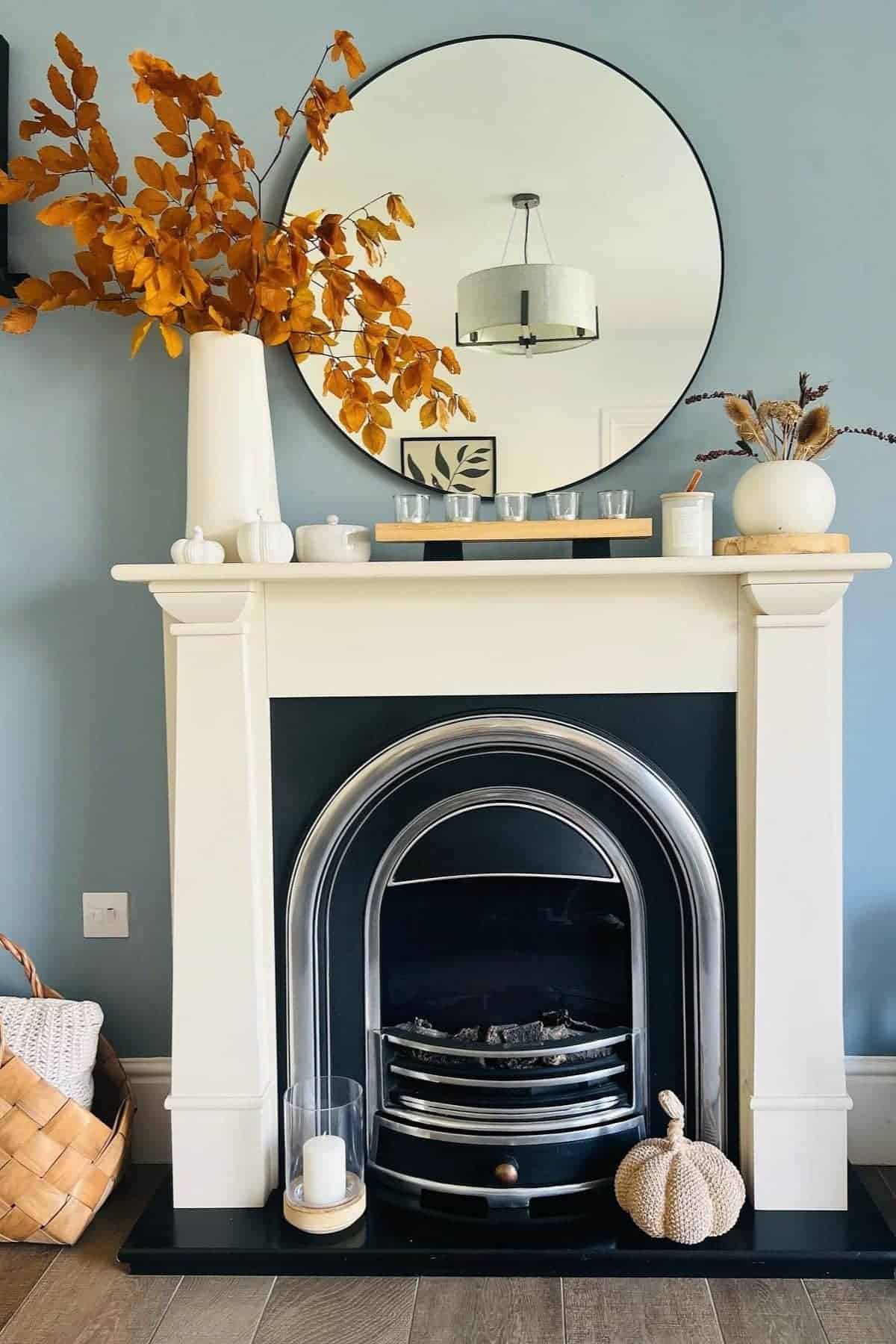 Modern fireplace with black and white surround, circular mirror above, autumnal vase arrangement, decorative pumpkins, candles, and wicker basket on wooden floor.