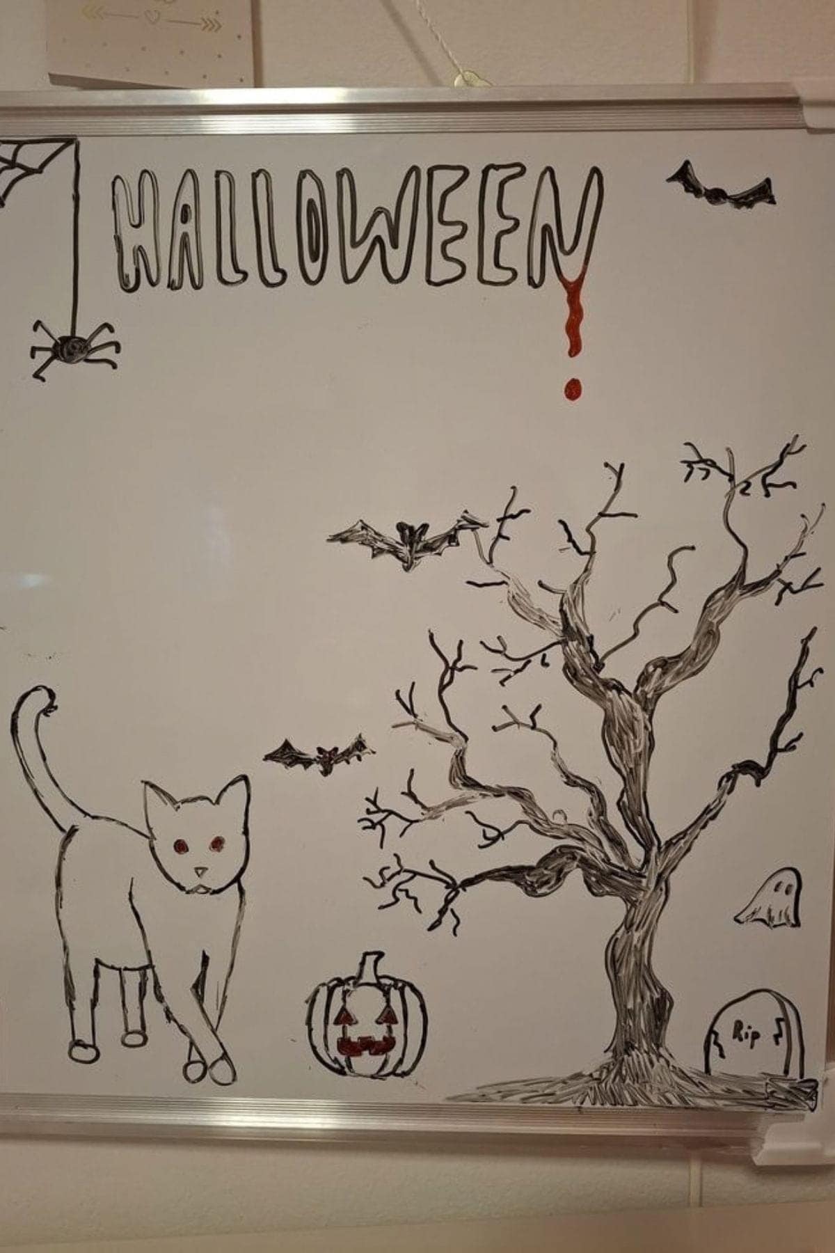 A whiteboard drawing of a cat, leafless tree, bats, spider, ghost, gravestone, and jack-o’-lantern beneath the word “HALLOWEEN” written in dripping letters.