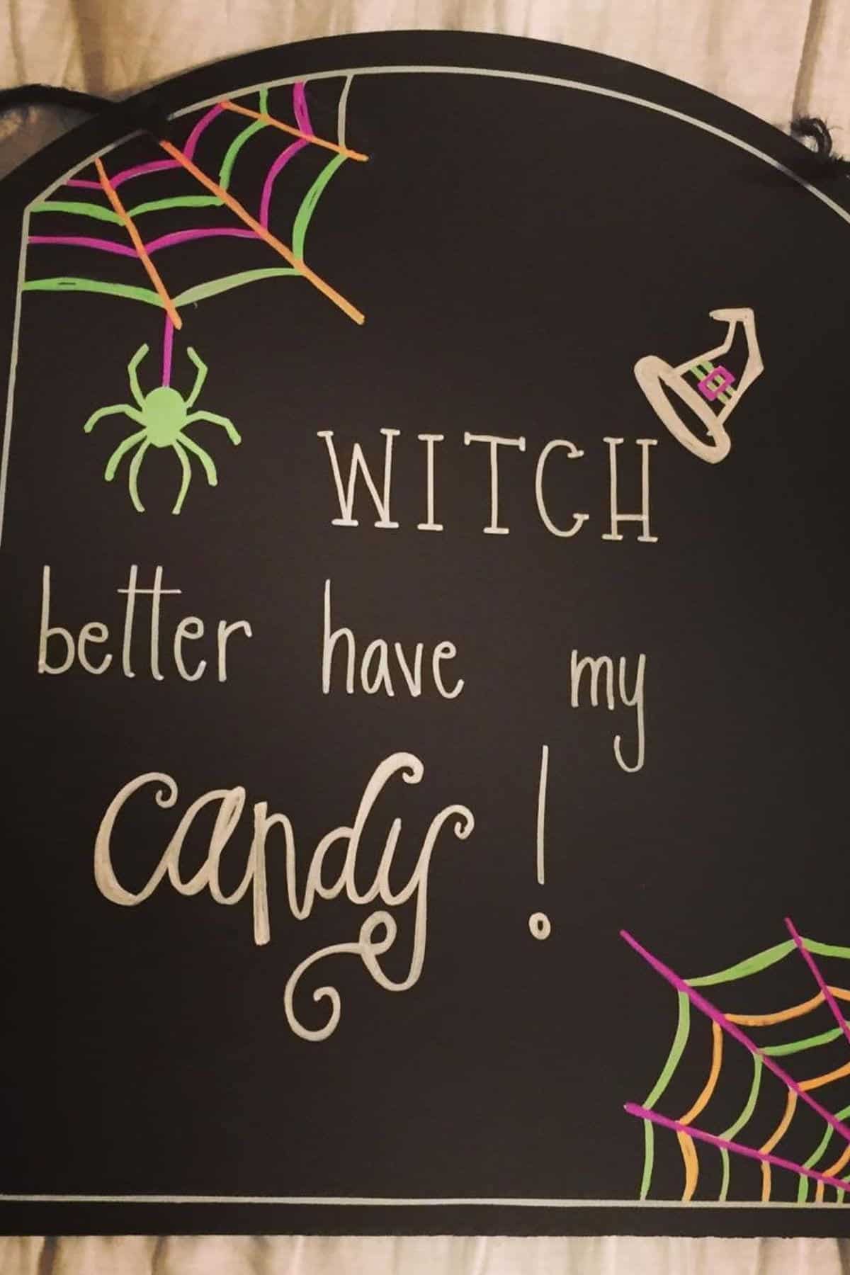 Black sign with colorful spider webs, a green spider, a witch hat, and the text “WITCH better have my candy!” written in playful fonts.