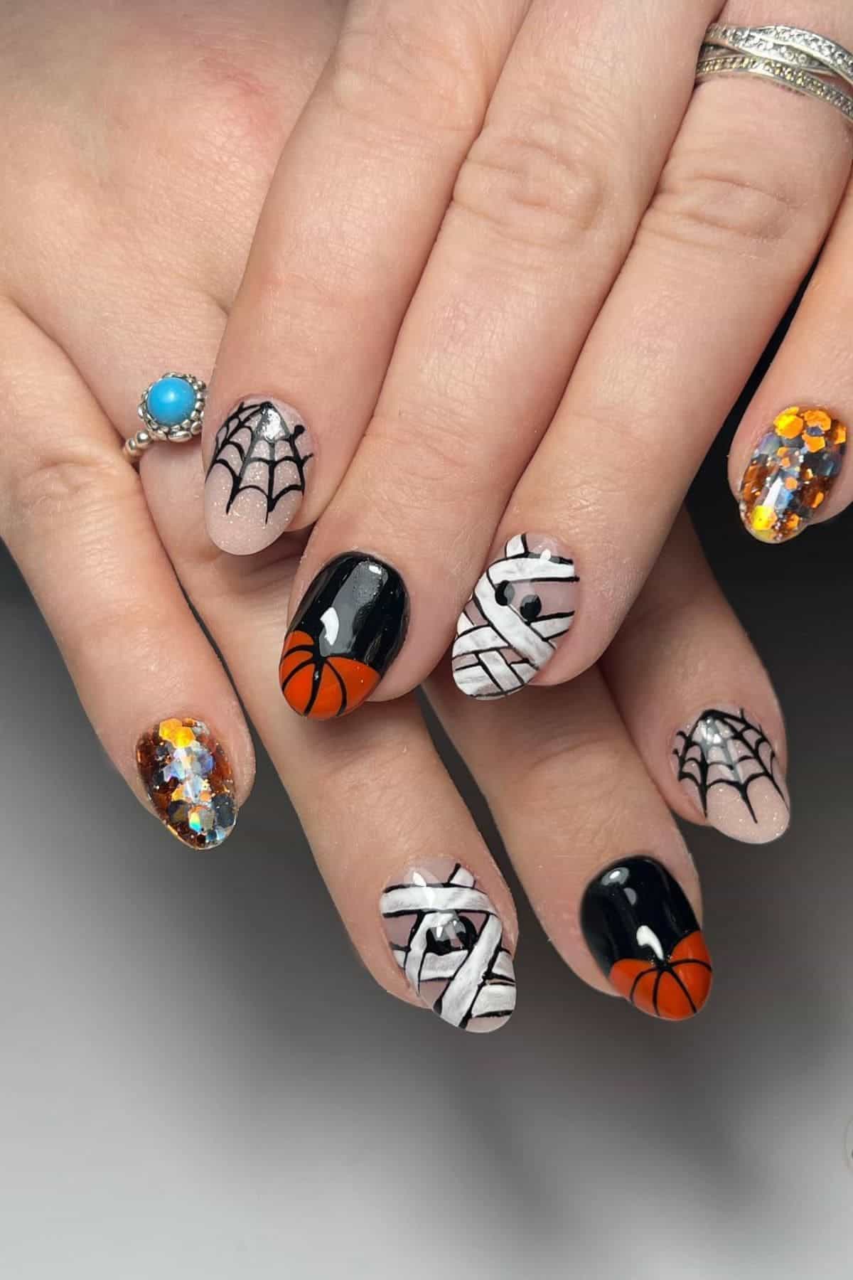 Hands with Halloween-themed nail art featuring spider webs, mummies, basketballs, and nails with orange and silver foil accents. The person wears a silver ring with a turquoise stone.