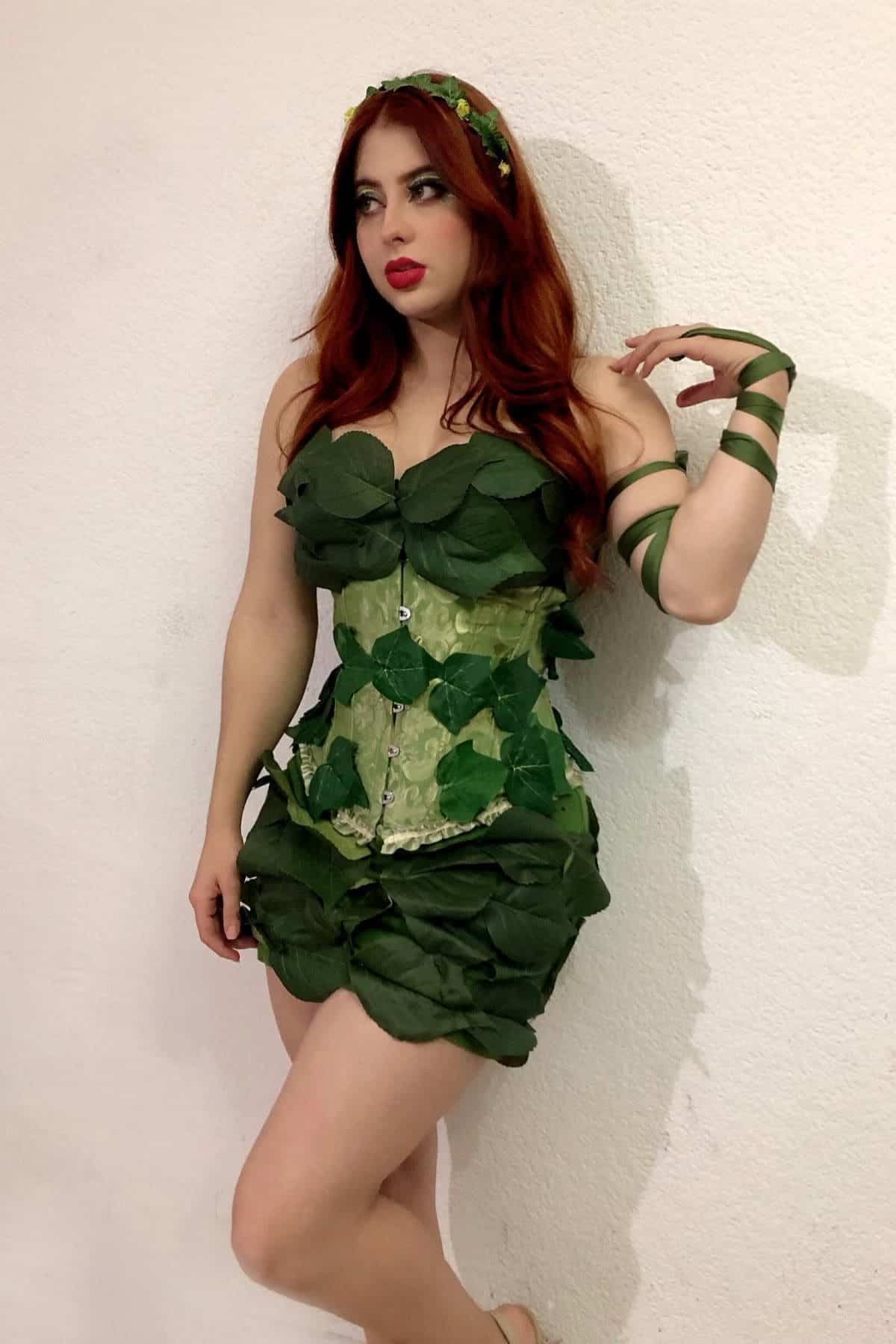 Person wearing a green leafy costume and headband, standing against a white wall with one arm wrapped in green ribbon.