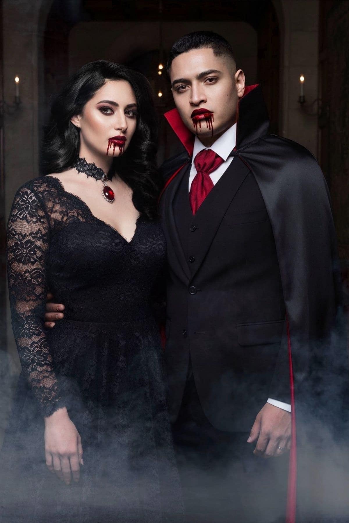 A woman and man dressed as vampires stand together, both with blood on their mouths, wearing black and red costumes, with mist at their feet.