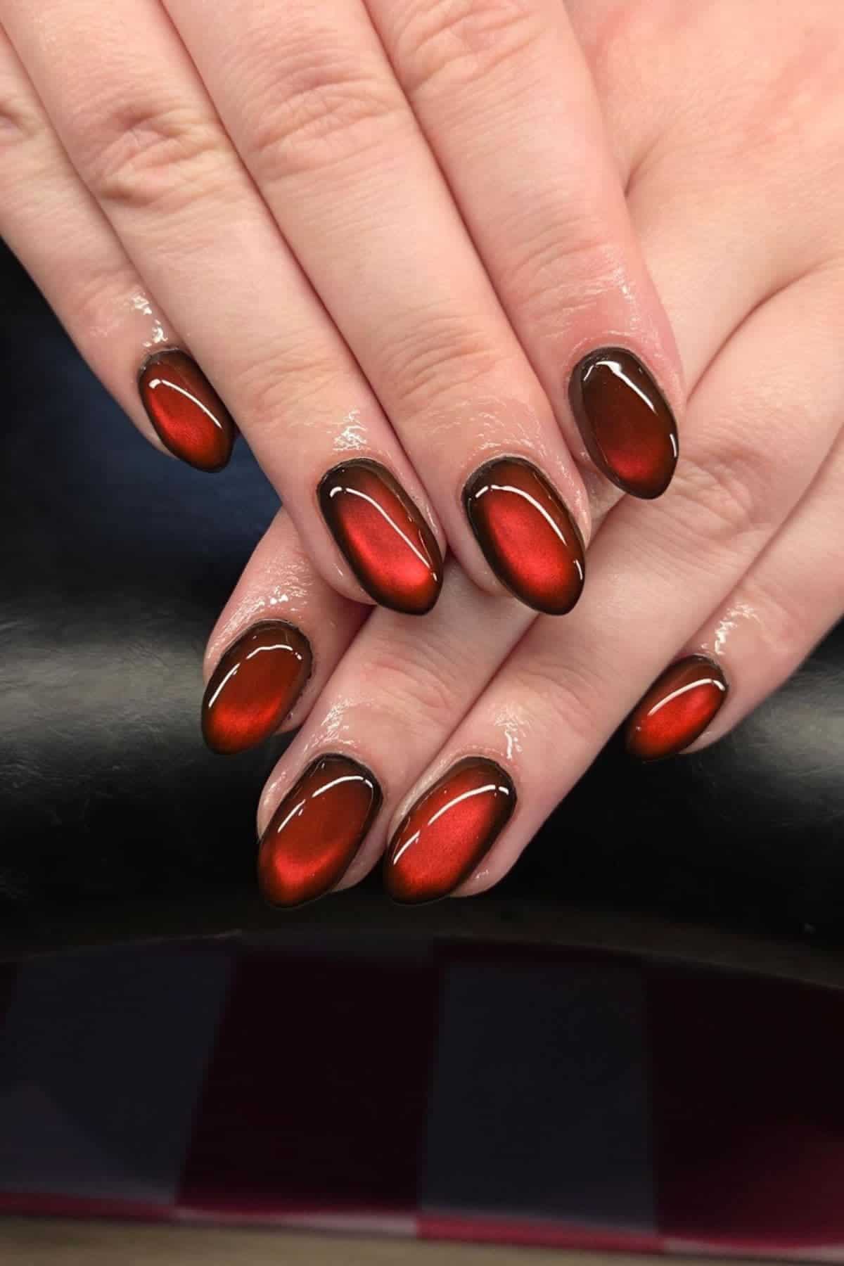 Close-up of hands with almond-shaped nails painted in a glossy, metallic red and black gradient design.