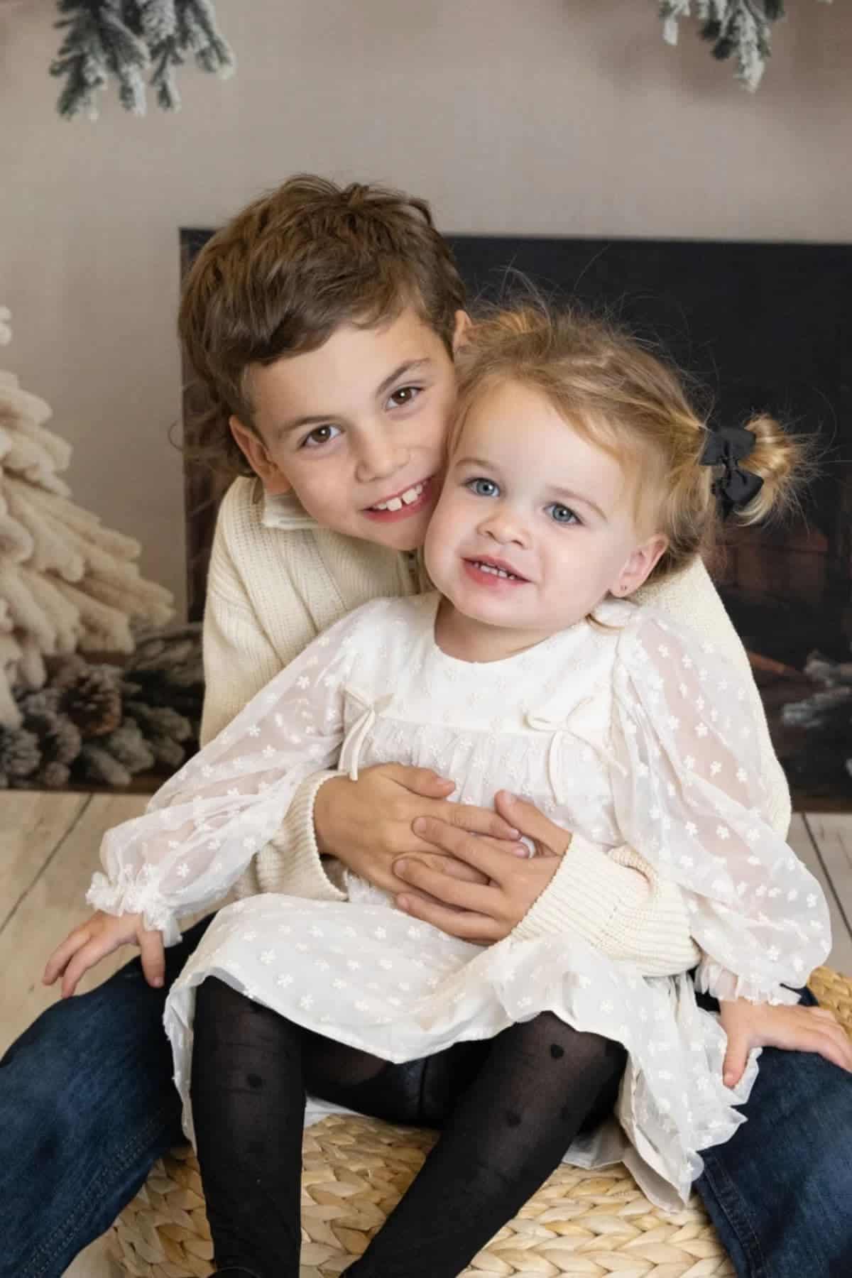 A young boy in a cream sweater sits behind a toddler girl in a white dress with black tights, smiling at the camera in a cozy, indoor holiday setting.