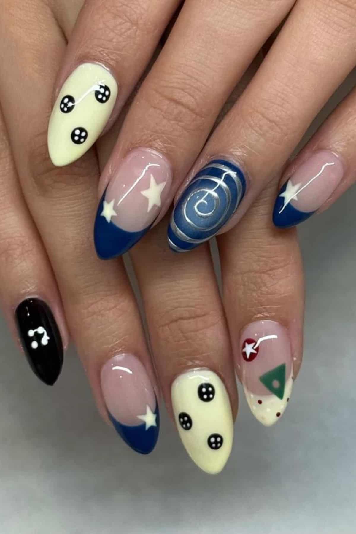 A hand with almond-shaped nails featuring a mix of blue, black, cream, and nude colors, decorated with stars, dots, buttons, and swirls.