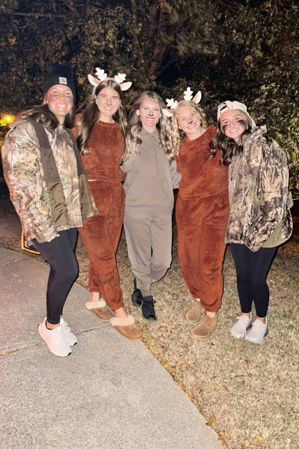 Five people stand outdoors at night, three in brown reindeer costumes and two in camouflage jackets, all smiling and posing for the photo.