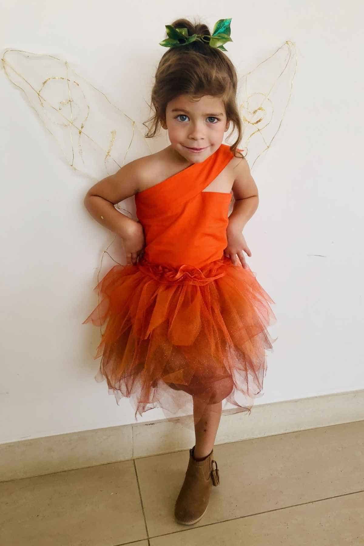 Child wearing an orange fairy costume with a tulle skirt, leafy hair accessory, and brown boots stands against a white wall with gold wire wings.