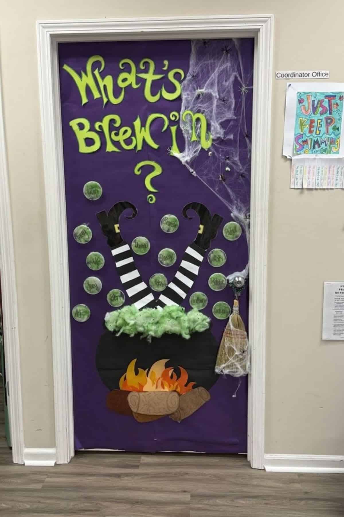 A door decorated with “What’s Brewin?” in green letters, witch legs in a cauldron, green bubbles, fake fire, cobwebs, and a broom, set in a white hallway.