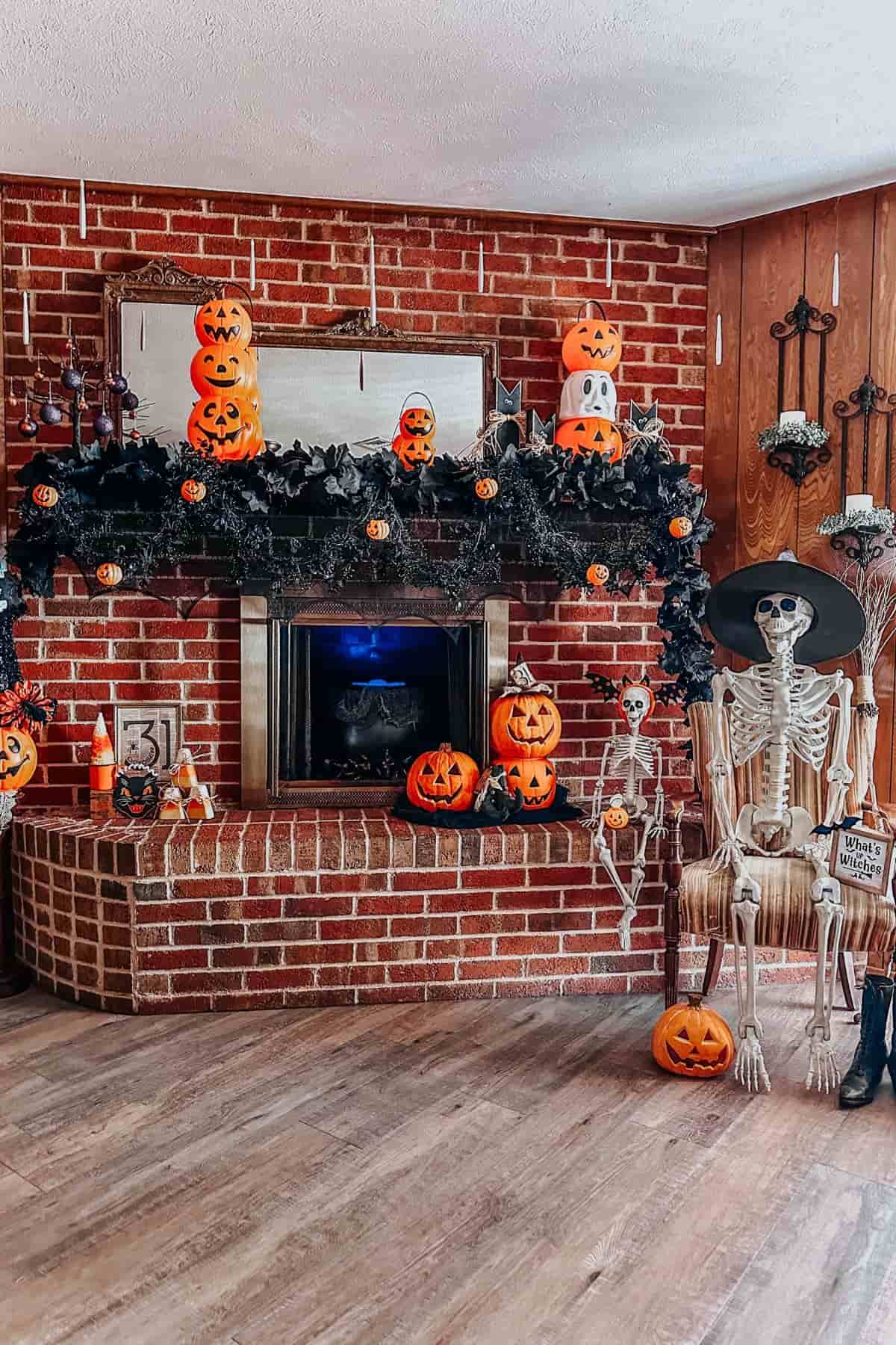 A brick fireplace decorated with jack-o’-lanterns, black garland, skeletons, and Halloween props. A skeleton sits on a chair beside pumpkins.
