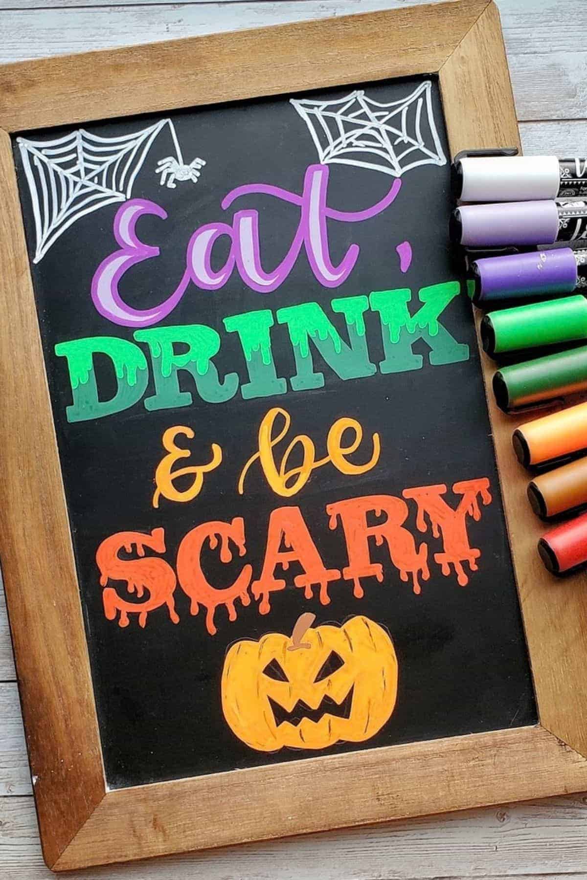 Chalkboard sign with “Eat, Drink & be Scary” in colorful letters, two spider webs, and a pumpkin drawing; colorful markers are beside the board.