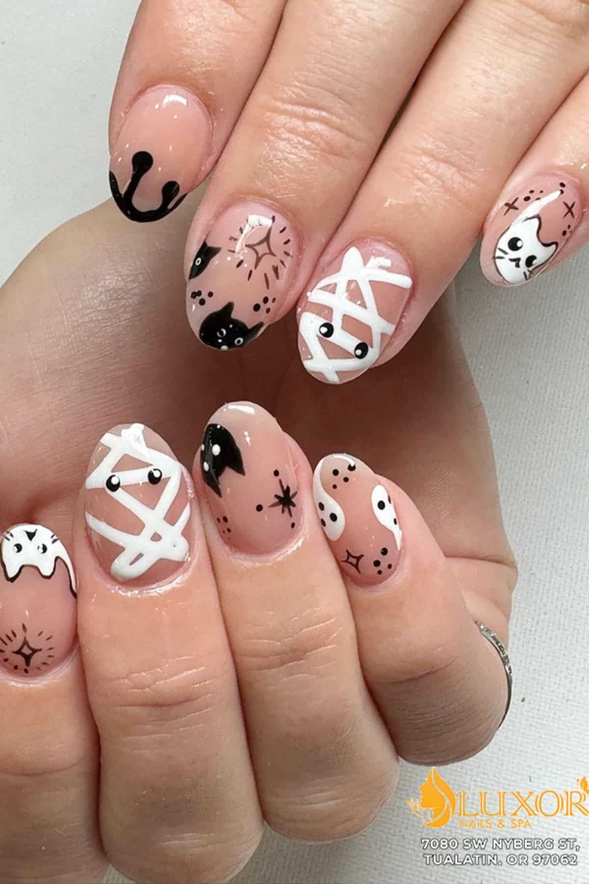 Hand with nails painted in Halloween-themed designs, including mummies, black cats, ghosts, and spider webs on a nude base.