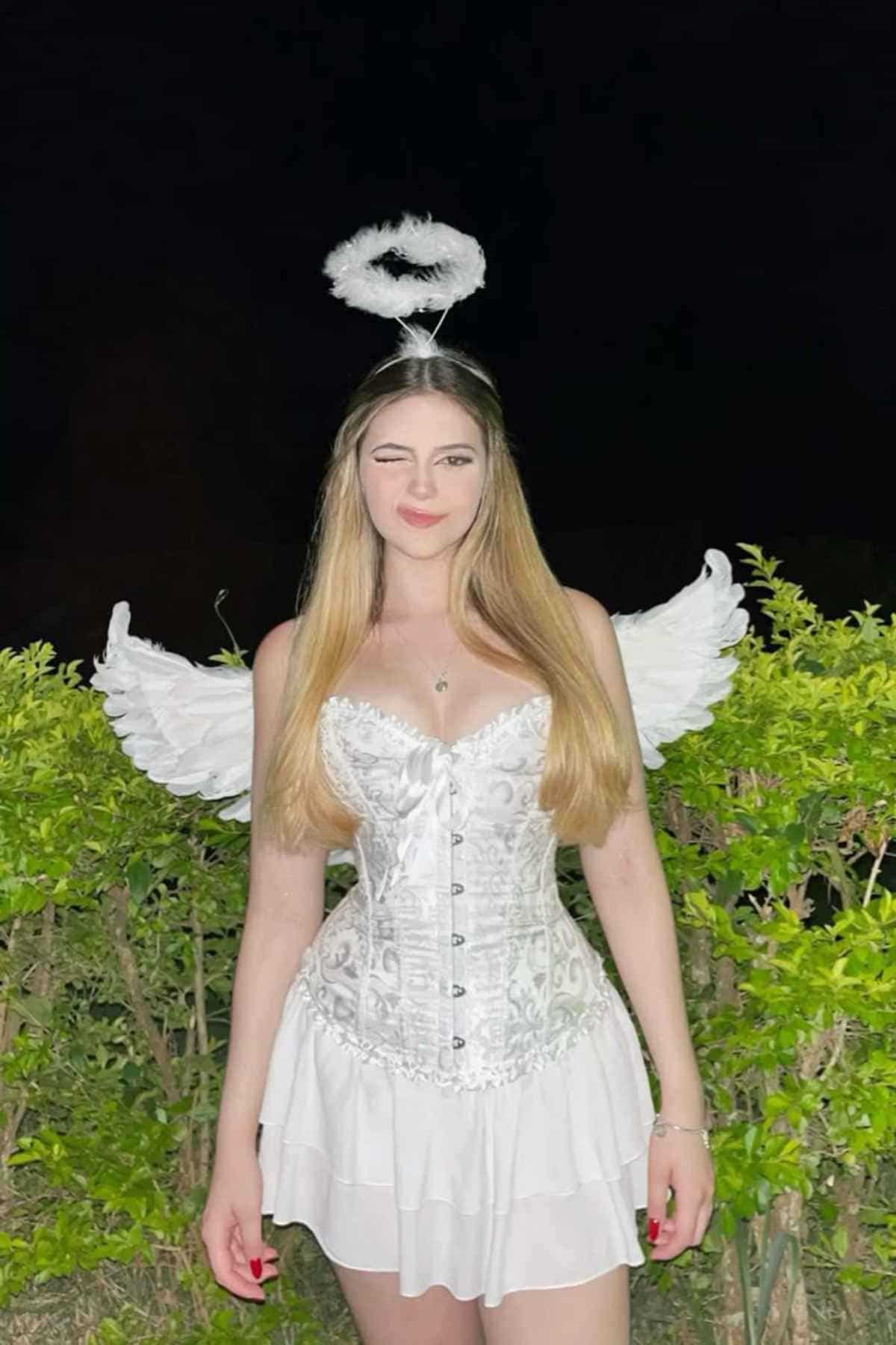 Woman dressed in an angel costume with wings and a halo stands in front of green bushes at night, winking at the camera.