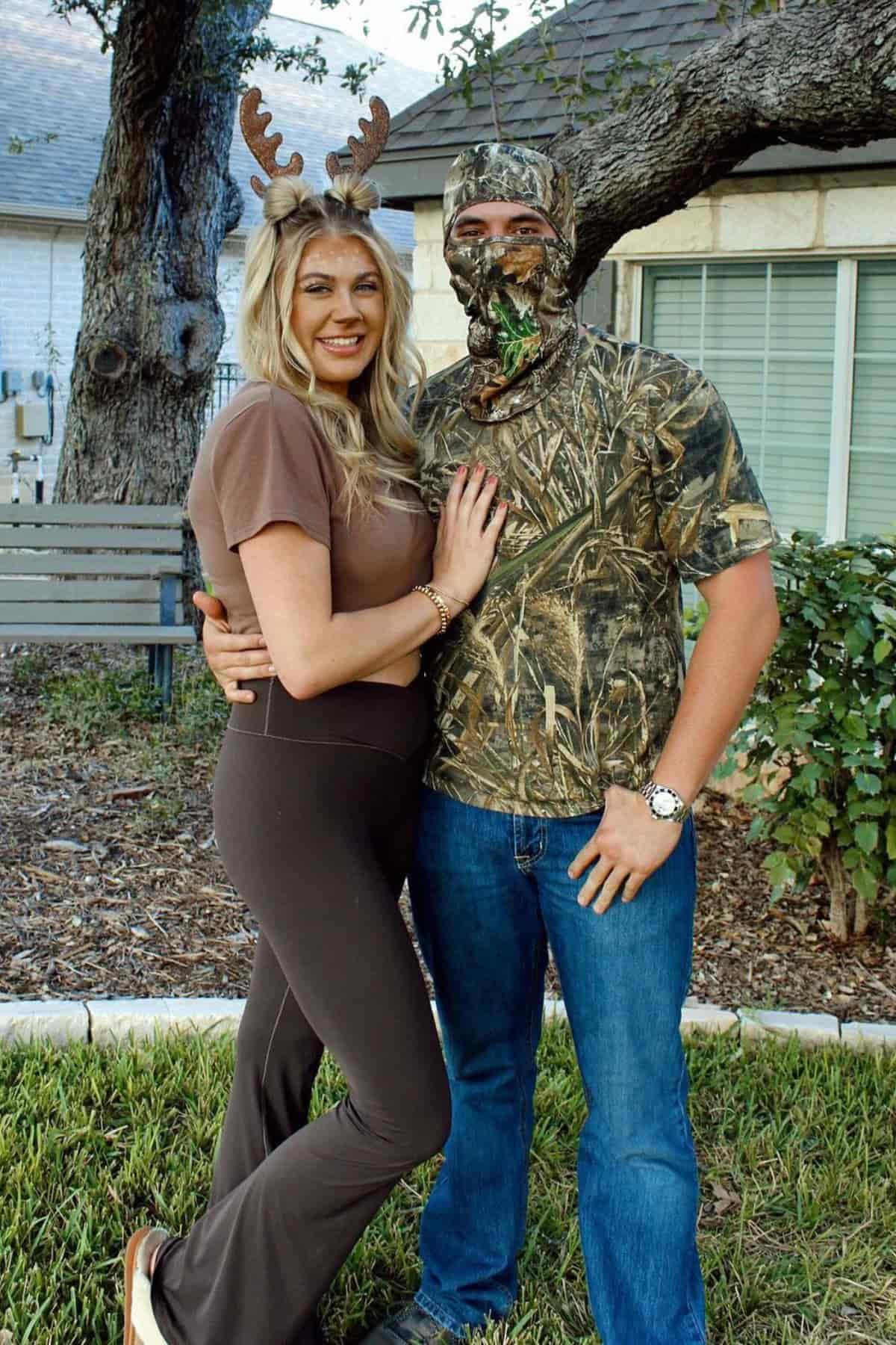 Woman wearing brown antlers and a man in camouflage clothing and a face covering pose together outside by a tree and a house.