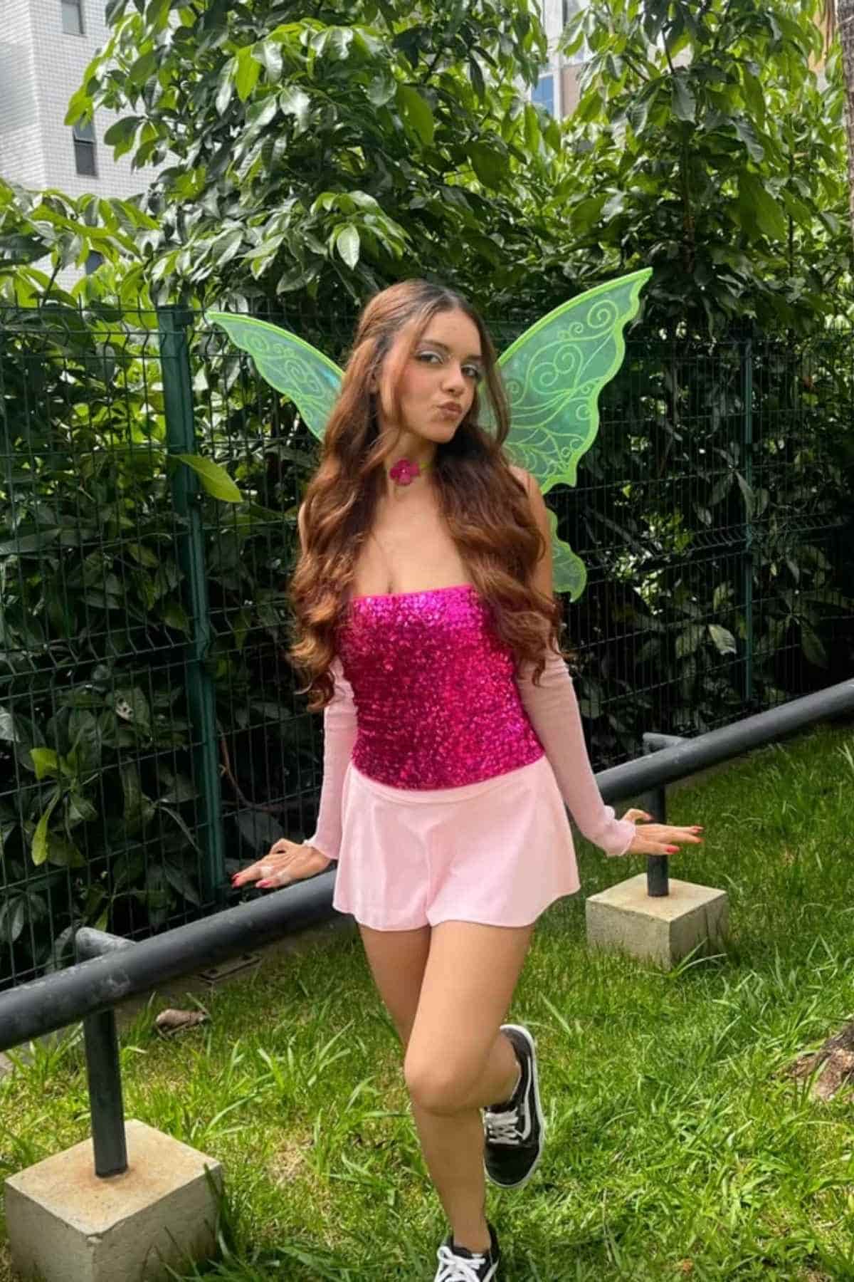 Woman wearing a pink sequin top, pink skirt, and green fairy wings poses outdoors near a fence and greenery.