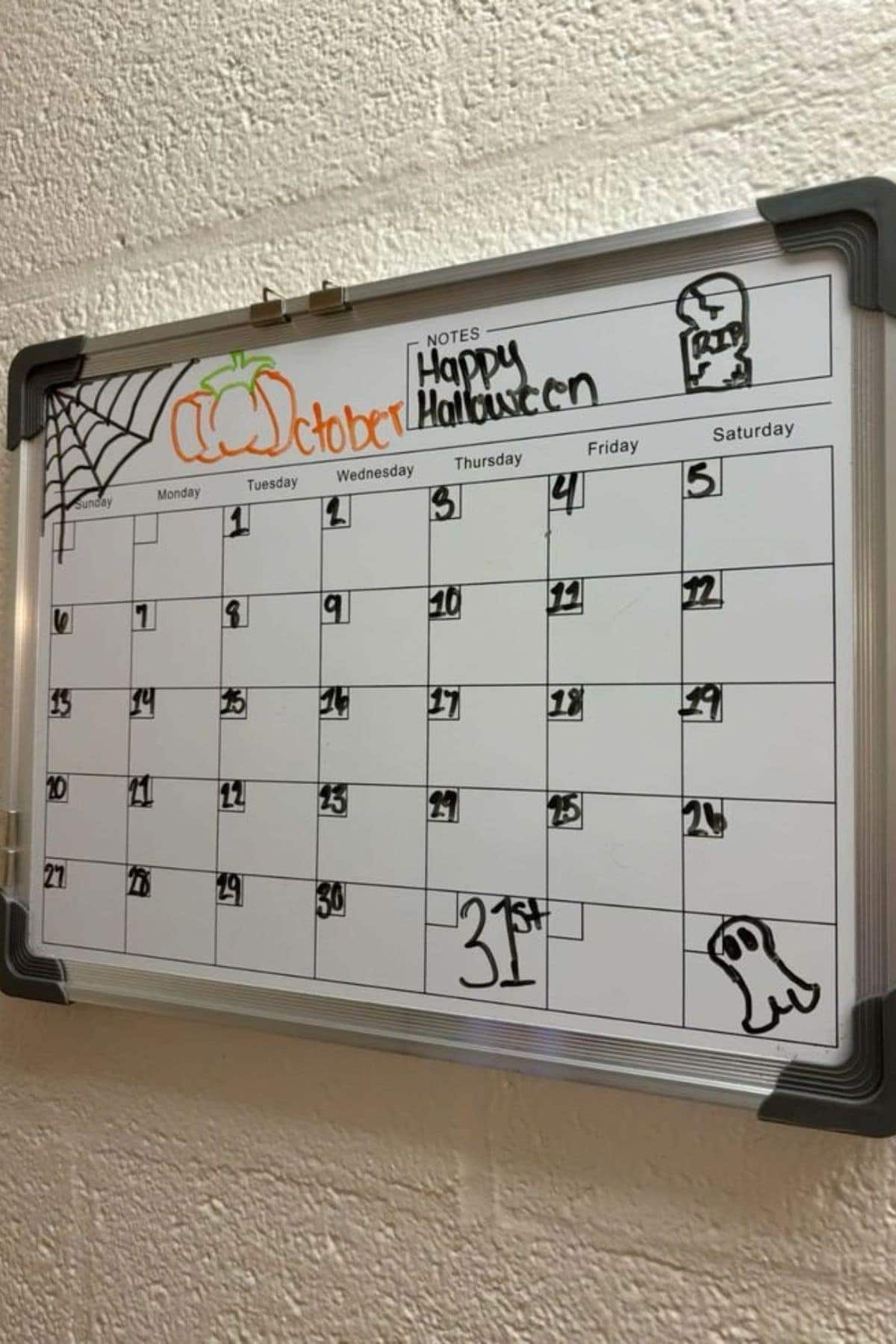 A whiteboard calendar for October features Halloween-themed doodles, including a pumpkin, spiderweb, ghost, and gravestone, with “Happy Halloween” written in the notes section.