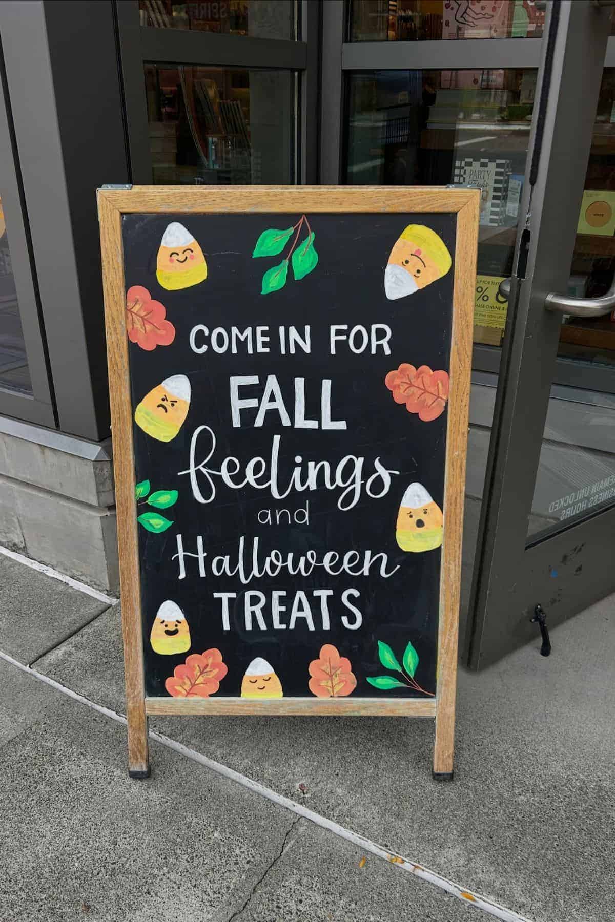 Chalkboard sign outside a store reads “Come in for Fall feelings and Halloween treats,” decorated with candy corn faces and autumn leaves.