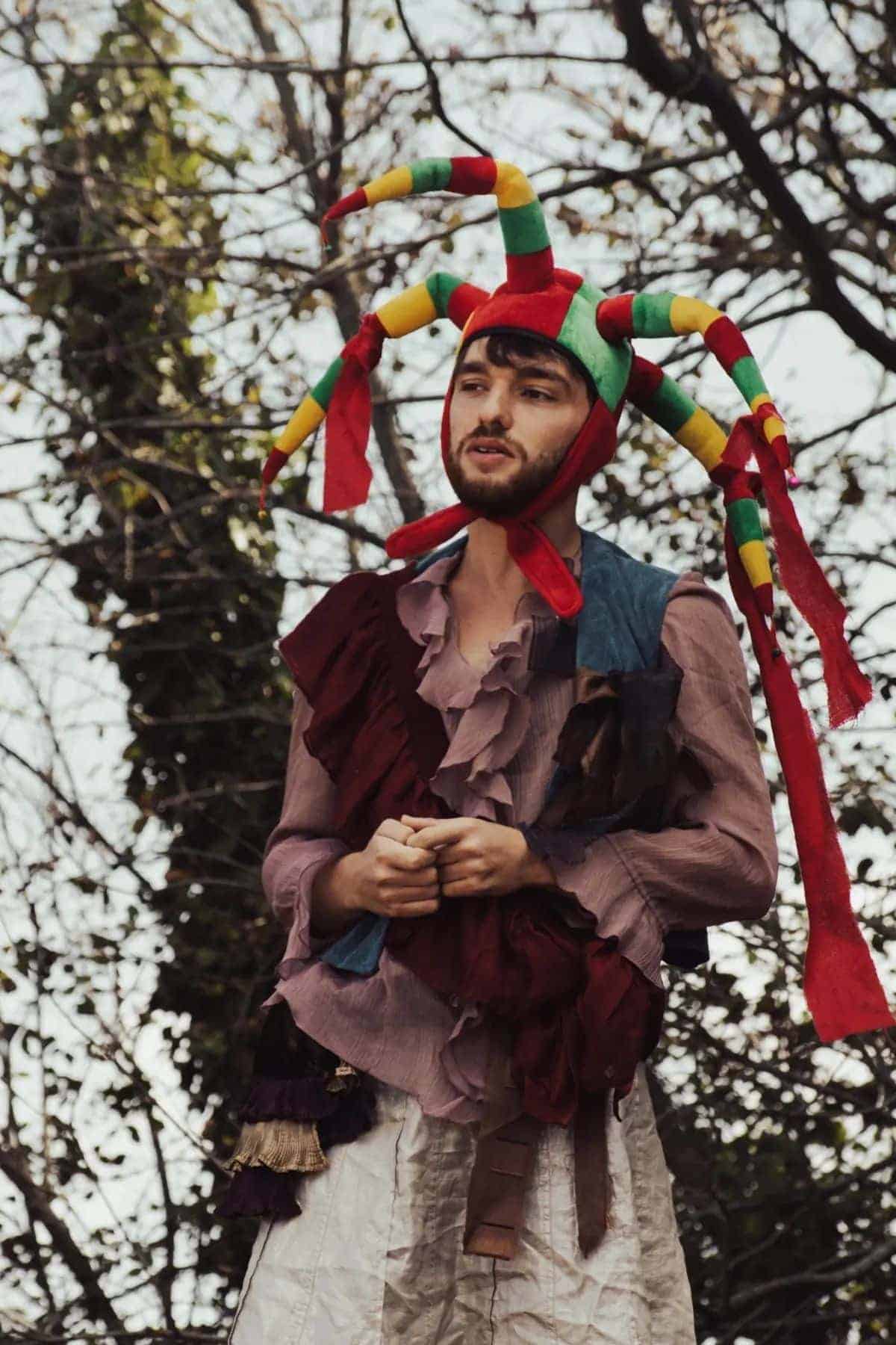 A person wearing a colorful jester hat and a patchwork costume stands outdoors in front of leafless trees.