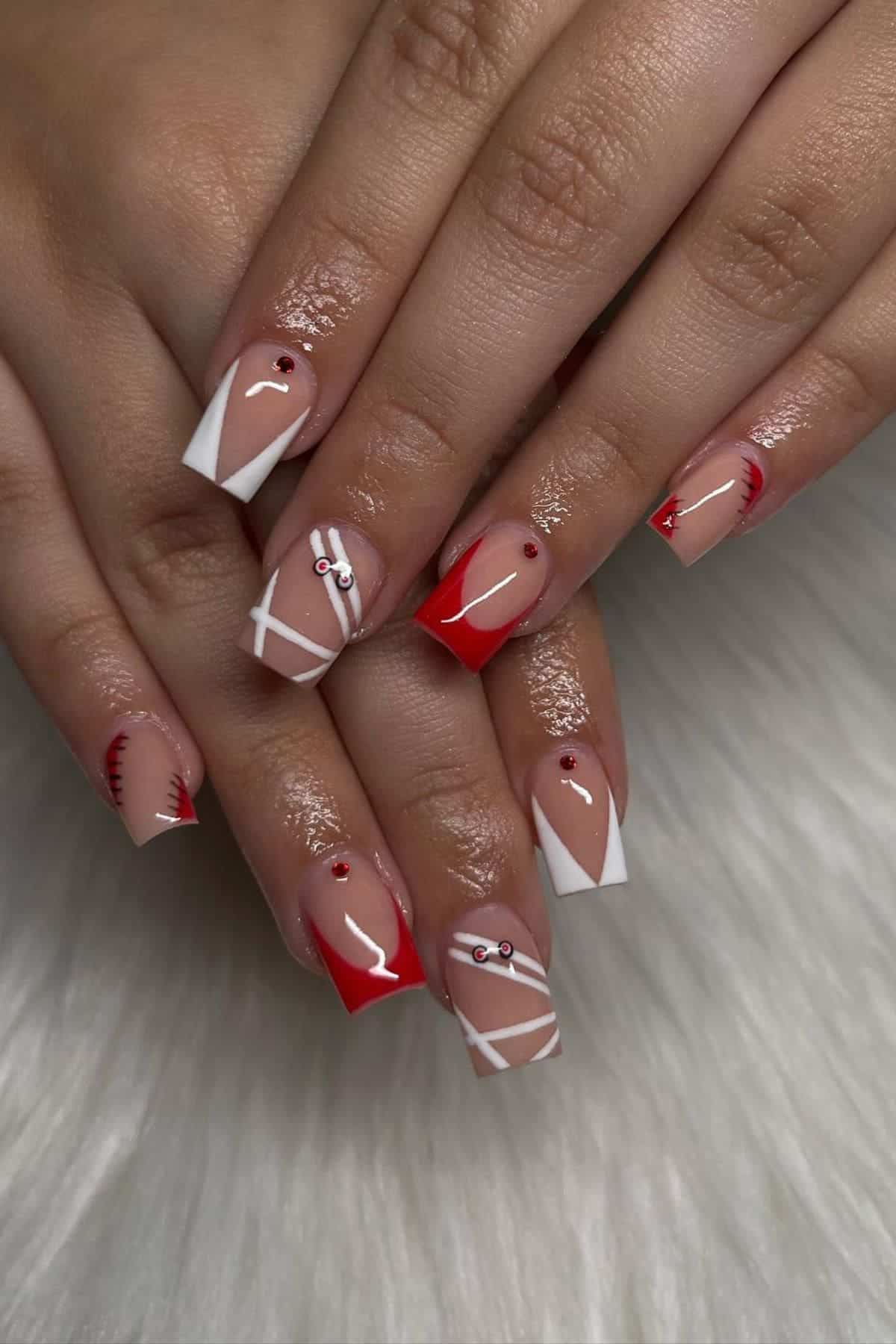 Hands with manicured square nails featuring red and white geometric designs, rhinestone accents, and a glossy finish on a white fur background.