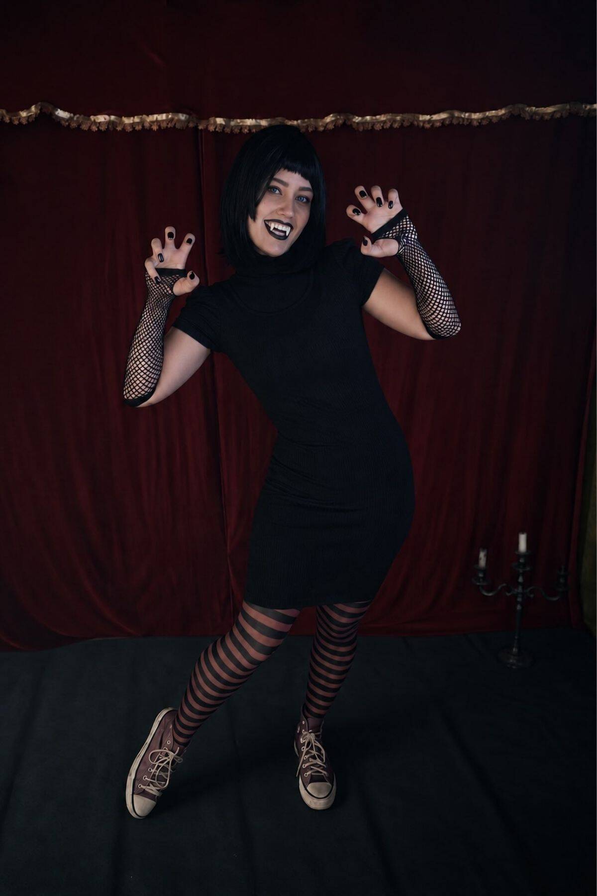 Person in black dress, striped tights, fishnet gloves, and black wig poses playfully with clawed hands against a red curtain backdrop. Candlestick holder is visible on the right.