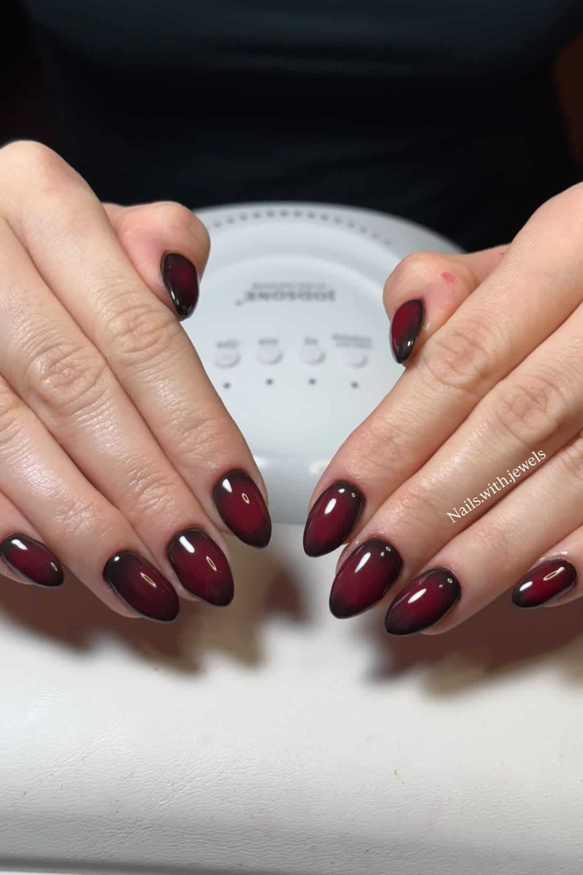 Hands with almond-shaped nails featuring a glossy dark red and black ombre manicure, resting on a white surface with a nail lamp in the background.