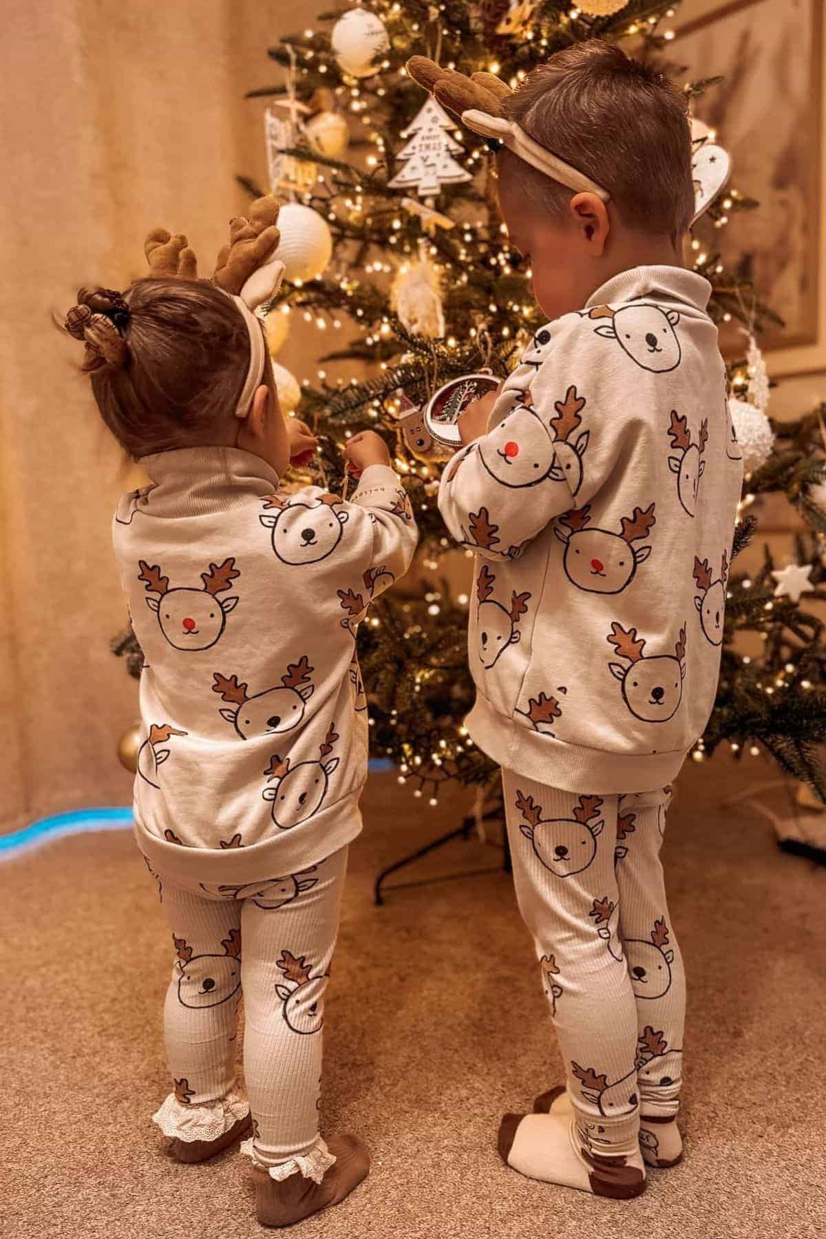 Two children wearing matching reindeer pajamas and antler headbands stand in front of a decorated Christmas tree.