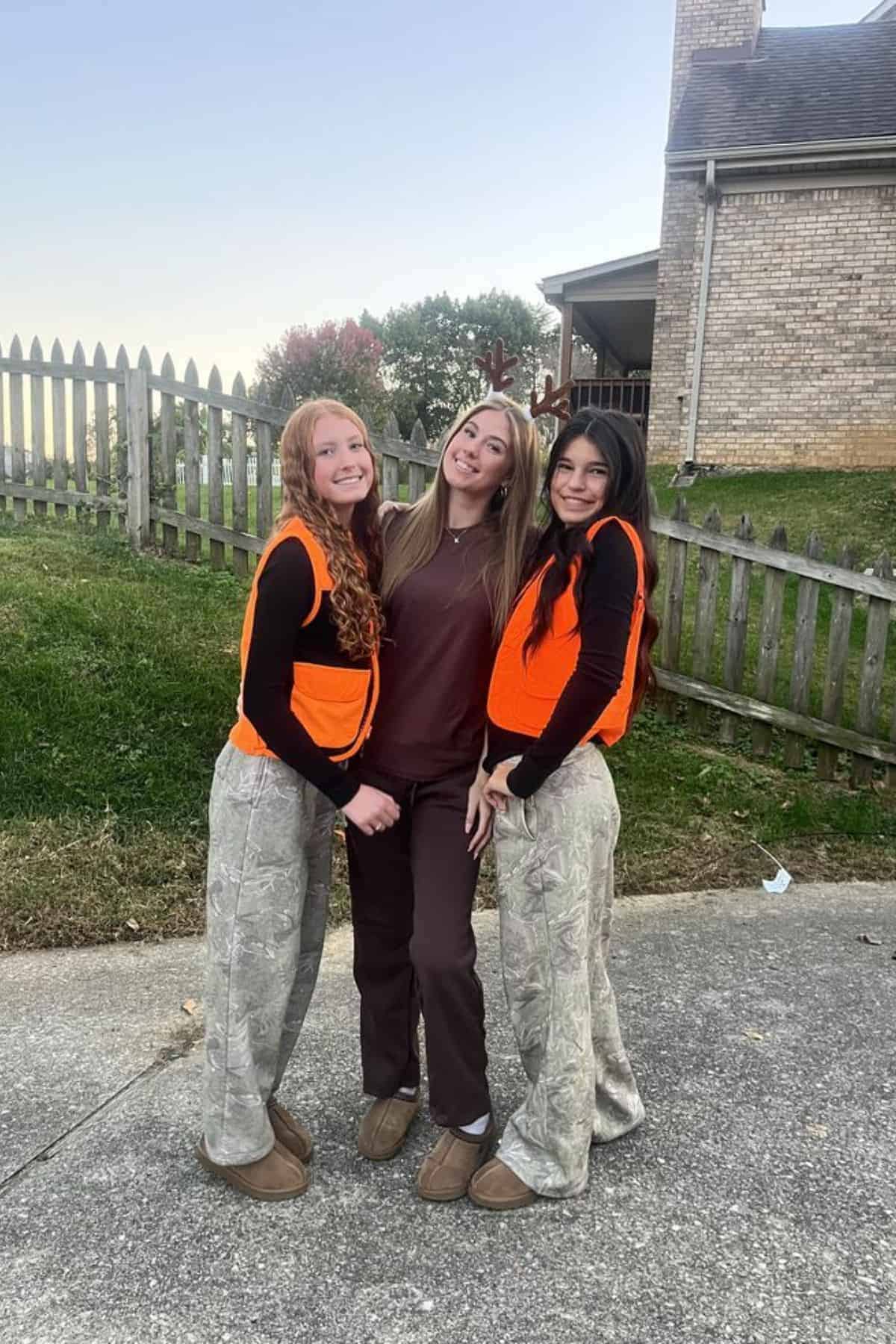 Three young women pose outdoors by a picket fence; two wear orange vests and camo pants, and one wears brown scrubs with antlers.