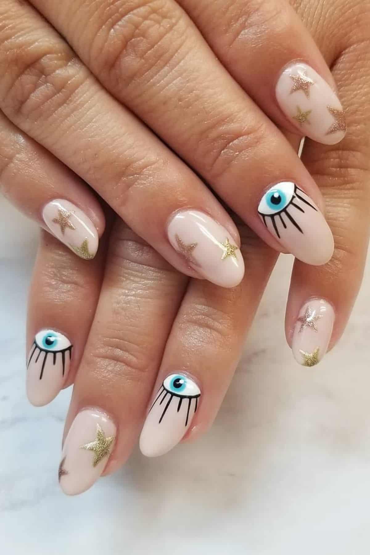 Hands with nude-colored almond nails featuring gold star designs and blue eye motifs with black lashes on accent nails.