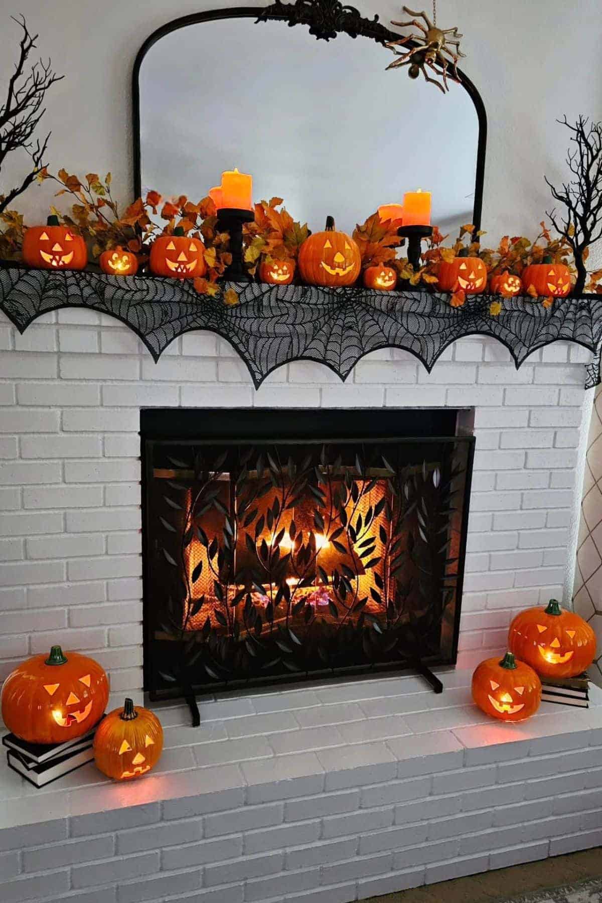 A white brick fireplace decorated with lit jack-o’-lanterns, fall leaves, black branches, and candles, with a fire burning behind a leaf-patterned screen.