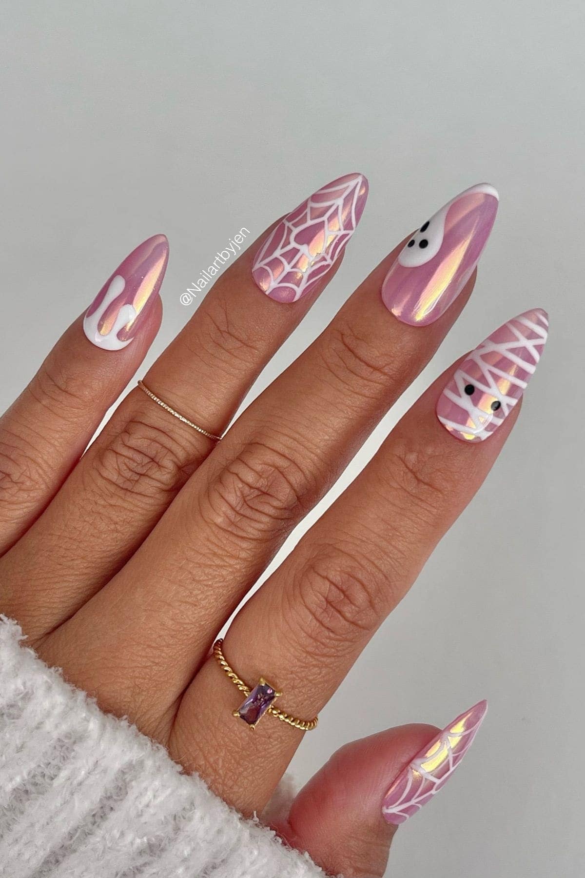 A hand with long, almond-shaped nails painted pink with white Halloween designs, including spider webs, a ghost, and a cat. The person wears two gold rings.