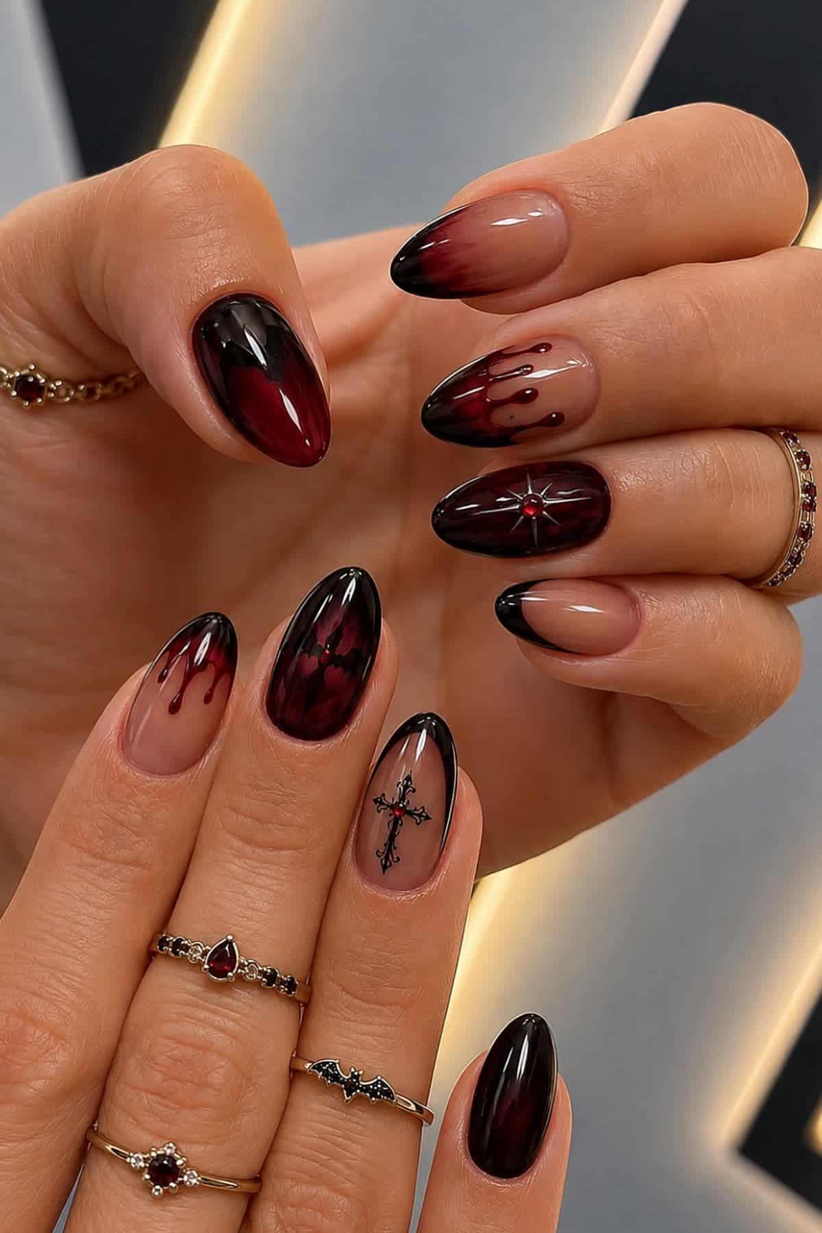 Hands with long almond-shaped nails featuring black and deep red ombre, drip designs, a star, and a cross accent. The person is wearing several delicate rings.