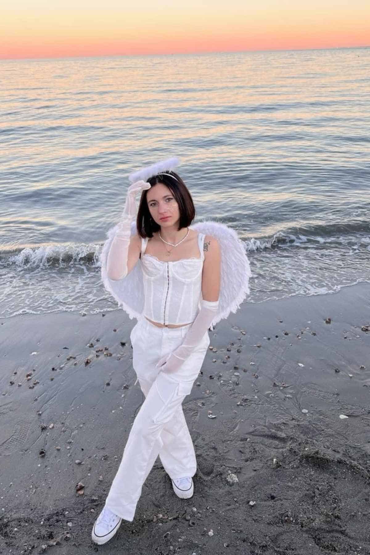 A person dressed in white angel costume stands on a beach near the shoreline at sunset, with calm waves and an orange sky in the background.