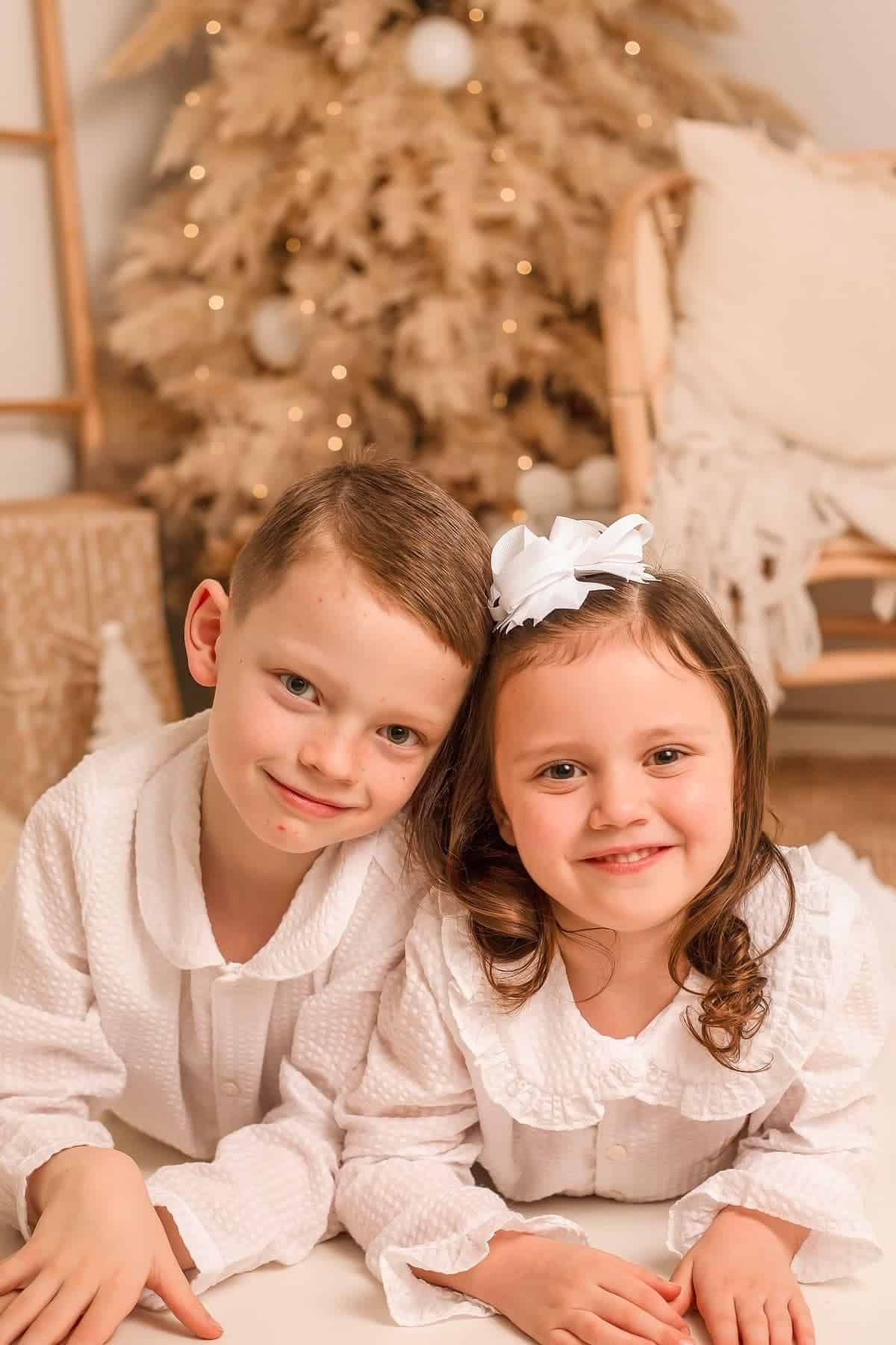 Two young children in white outfits smile at the camera with a decorated tree and cozy holiday decor in the background.