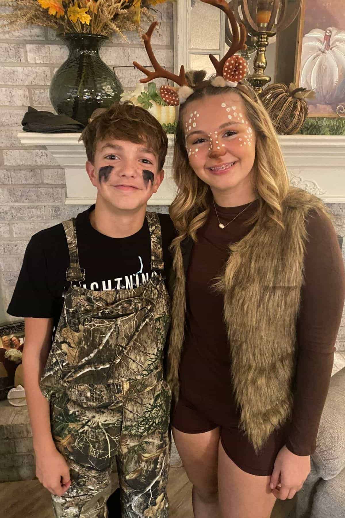 Two teens in costumes: one dressed as a hunter in camo overalls with face paint, the other as a deer with antlers and face makeup. They are standing indoors in front of a fireplace.