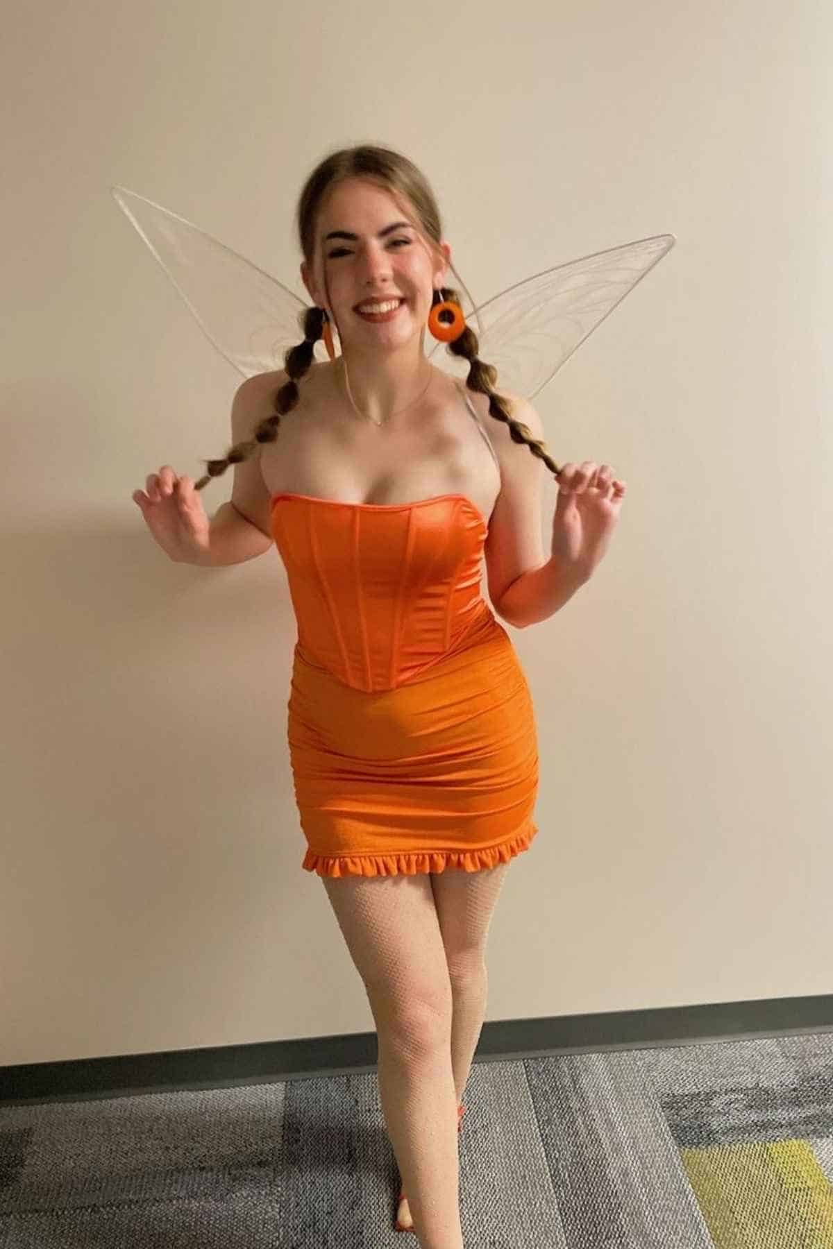 Person dressed in an orange strapless dress with wing accessories and braided hair, standing indoors on a patterned carpet.