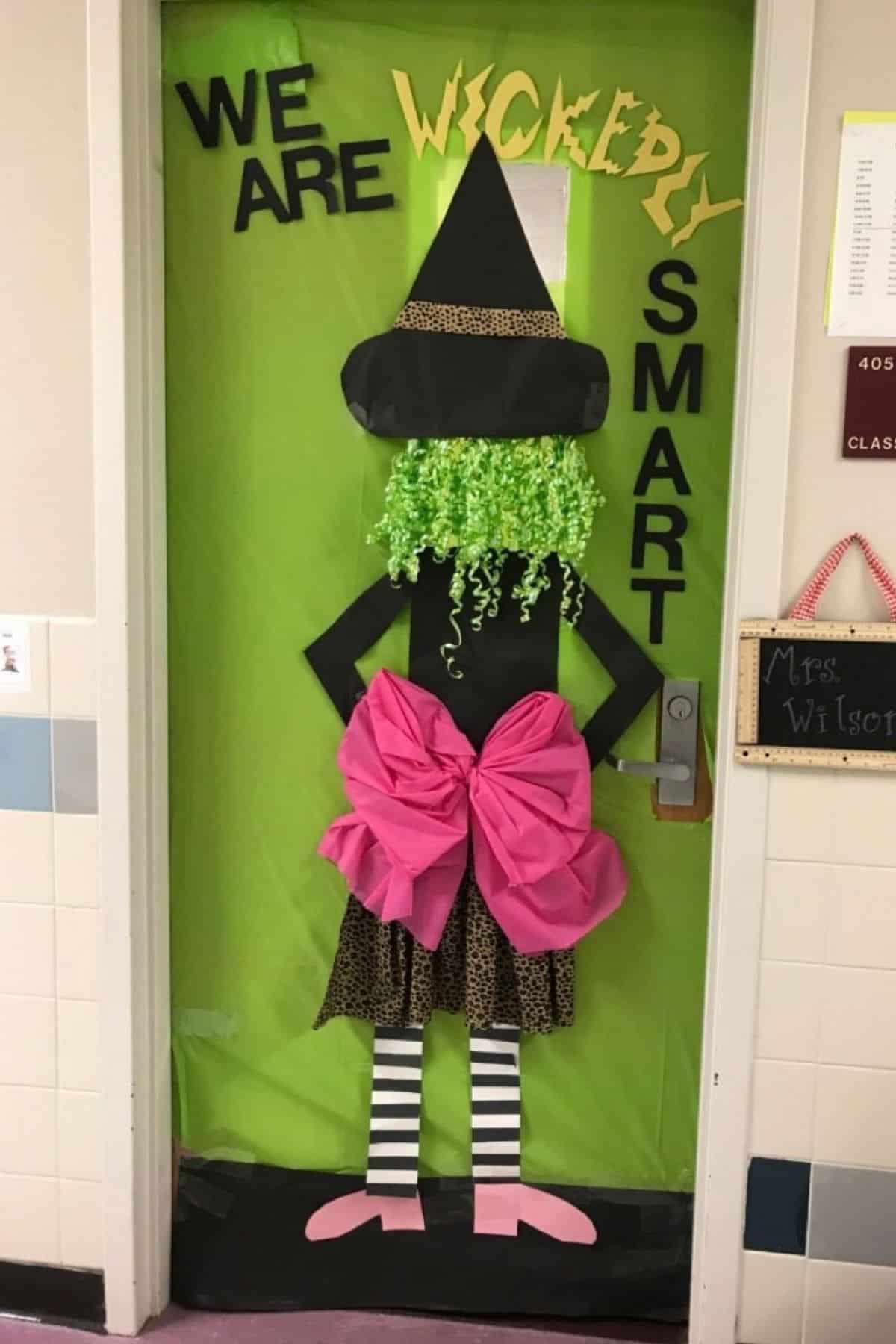 A classroom door decorated with a witch figure, green background, pink bow, and the words “We are wickedly smart” in large letters.