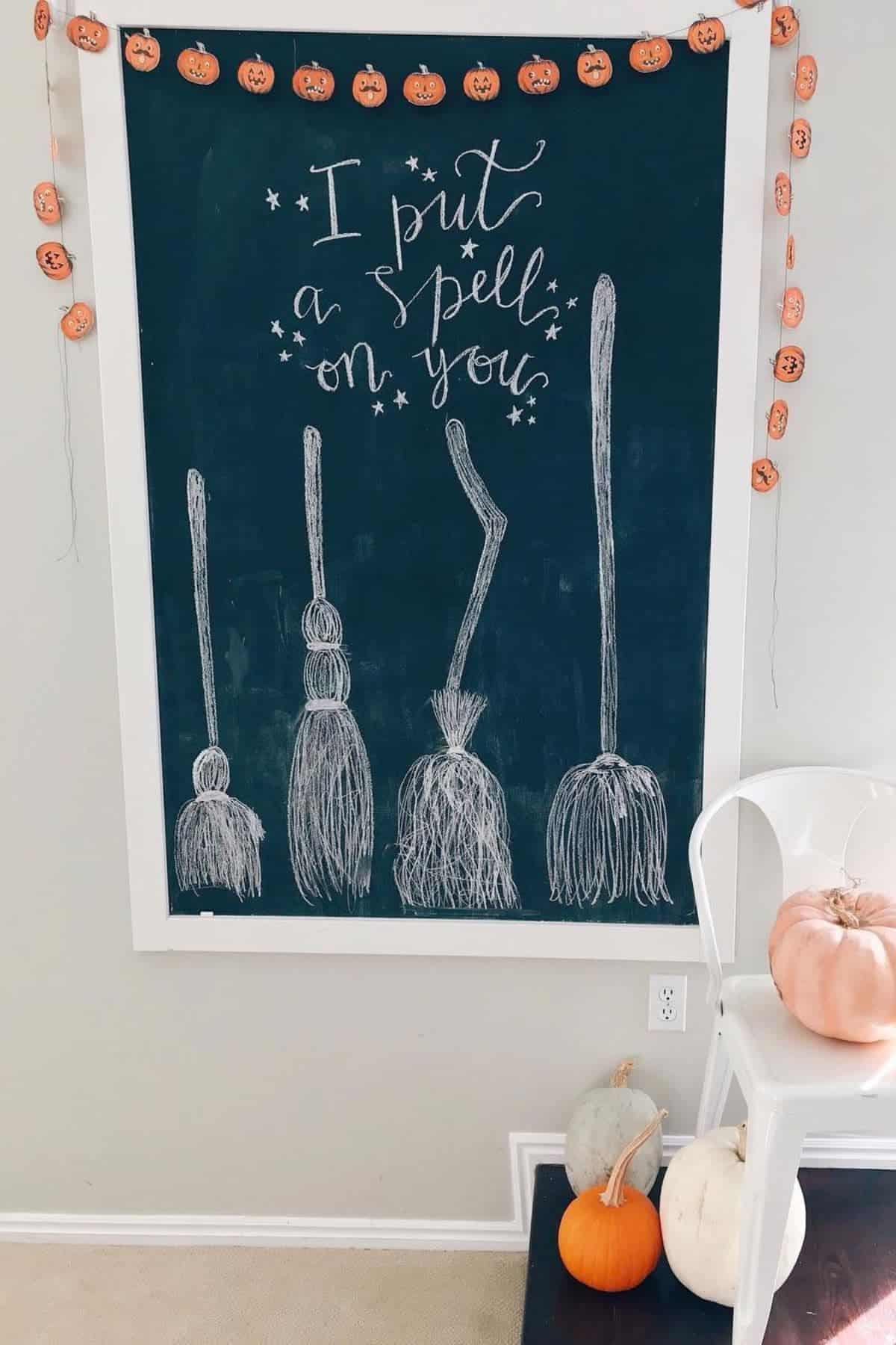 A chalkboard with “I put a spell on you” written above drawings of four brooms. Decorative garland, pumpkins, and a white chair are in the scene.