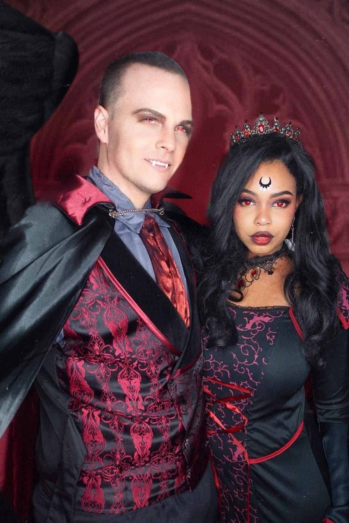 Two people in elaborate red and black gothic costumes pose together; one wears a cape, the other a crown and a crescent moon symbol on their forehead, both with red eye makeup.