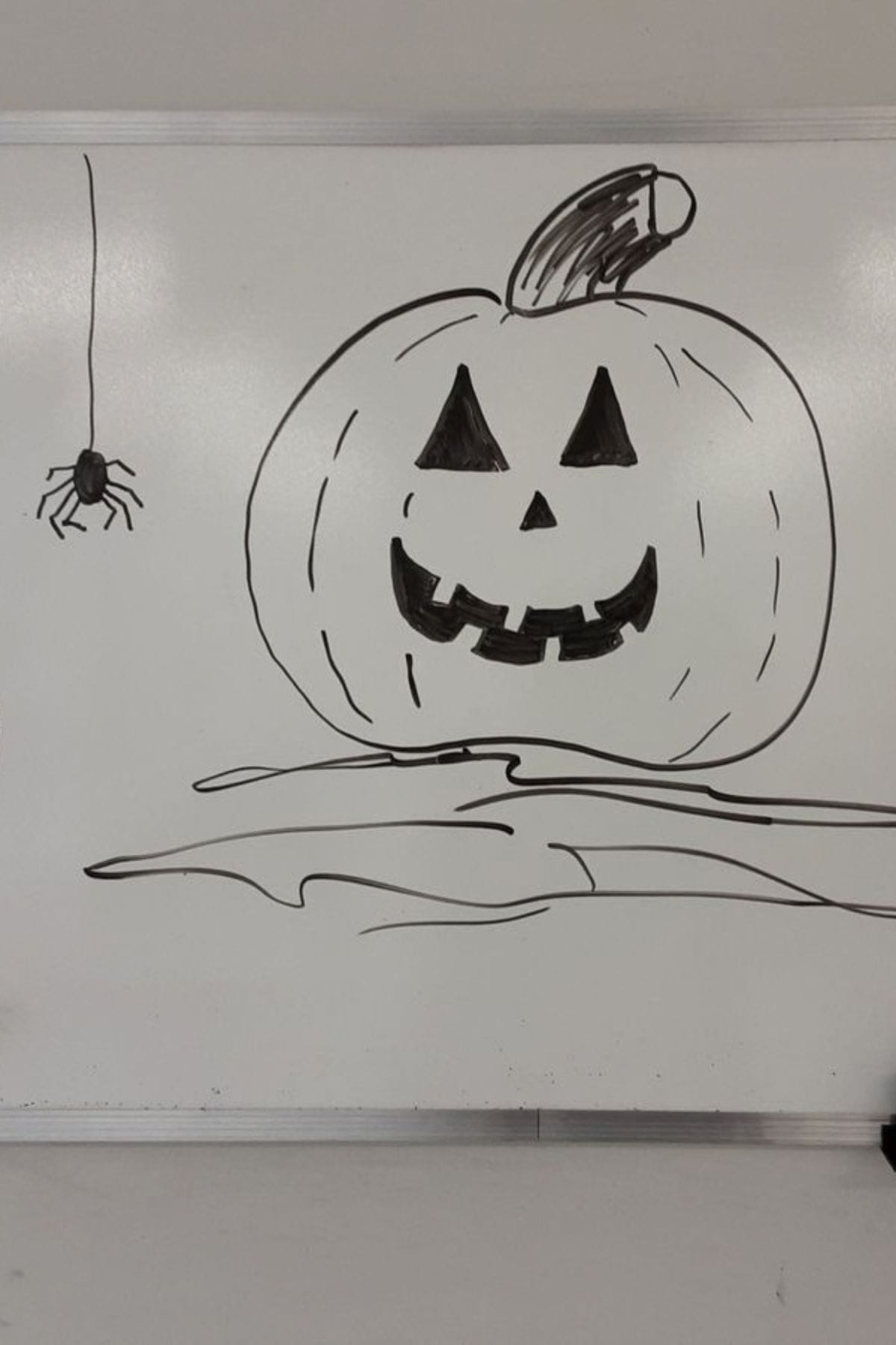Black marker drawing of a jack-o’-lantern with a smiling face on a whiteboard, accompanied by a hanging spider on the left side.