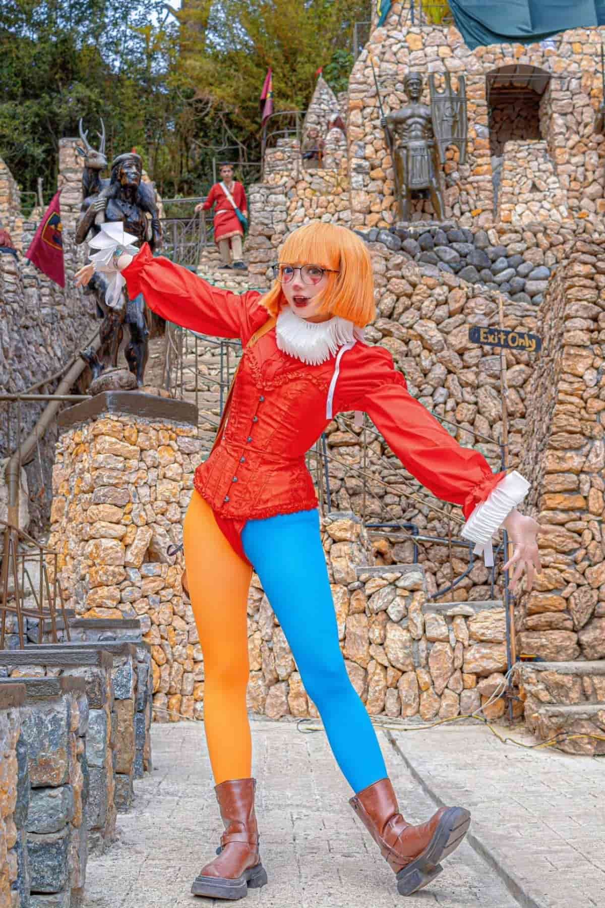 Person in a red and orange clown costume with blue and orange tights poses outdoors on a stone walkway, surrounded by stone structures and statues.