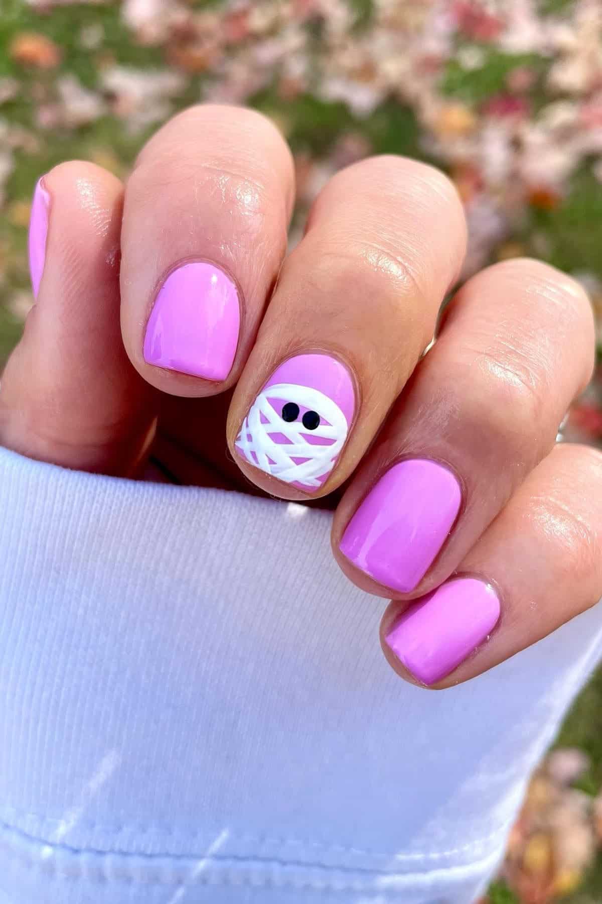 A hand with light purple nail polish; the ring fingernail has a white mummy design with two black eyes. The background is grass with scattered autumn leaves.